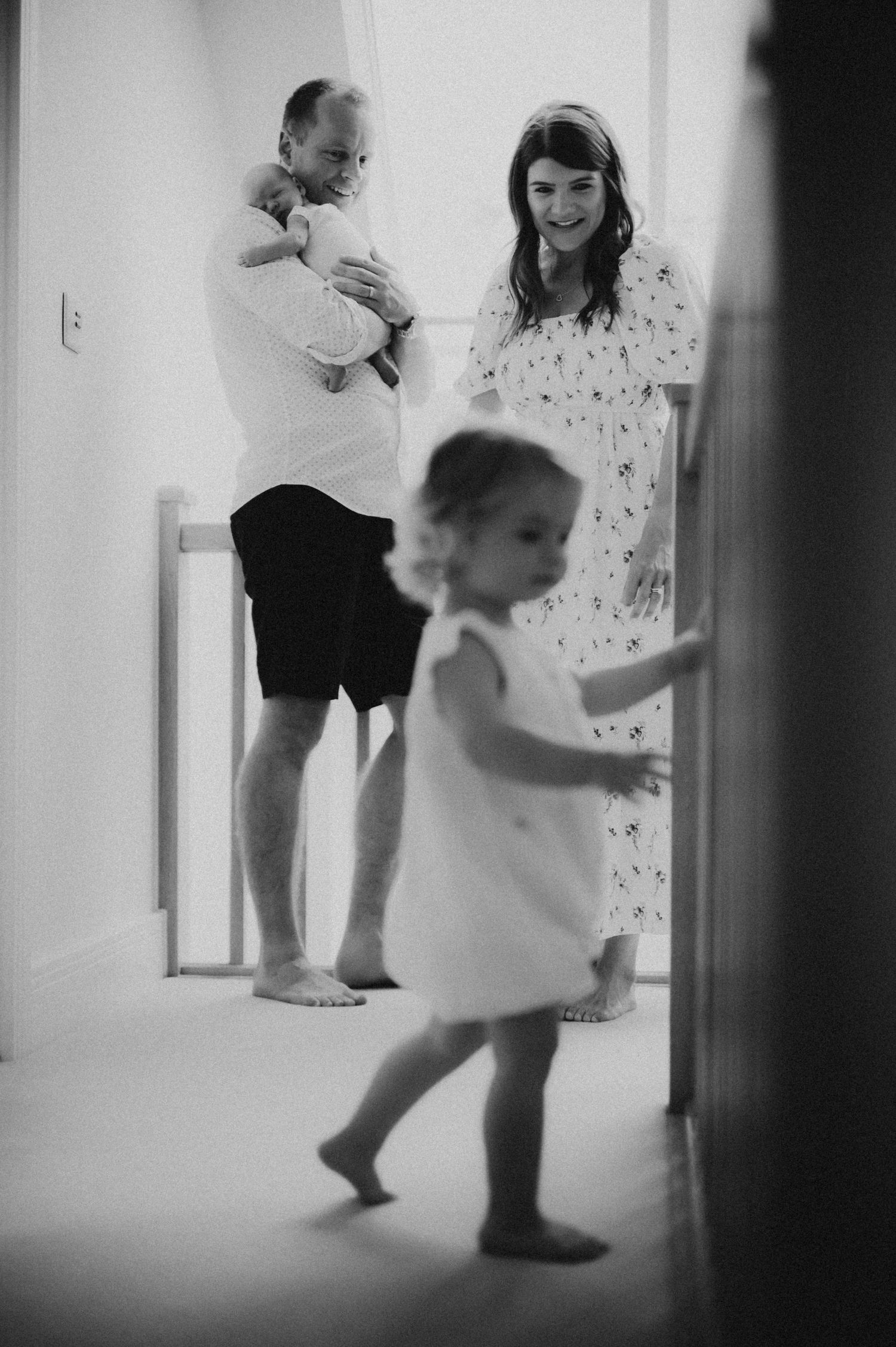 Black and white family photo of parents with newborn baby and toddler daughter walking in bright hallway.