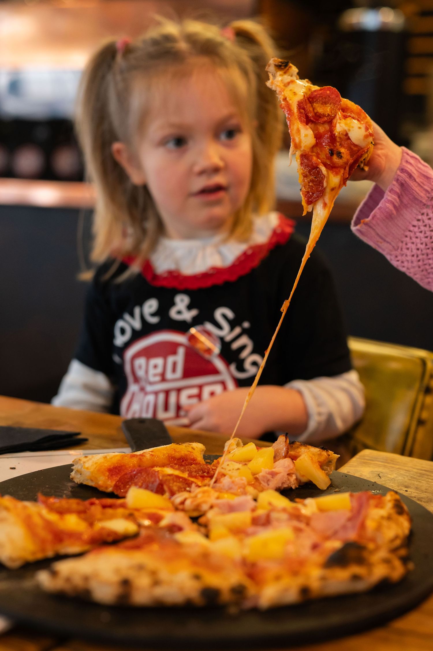A young girl watches as a cheesy slice of pizza is lifted from a stone plate at a restaurant.