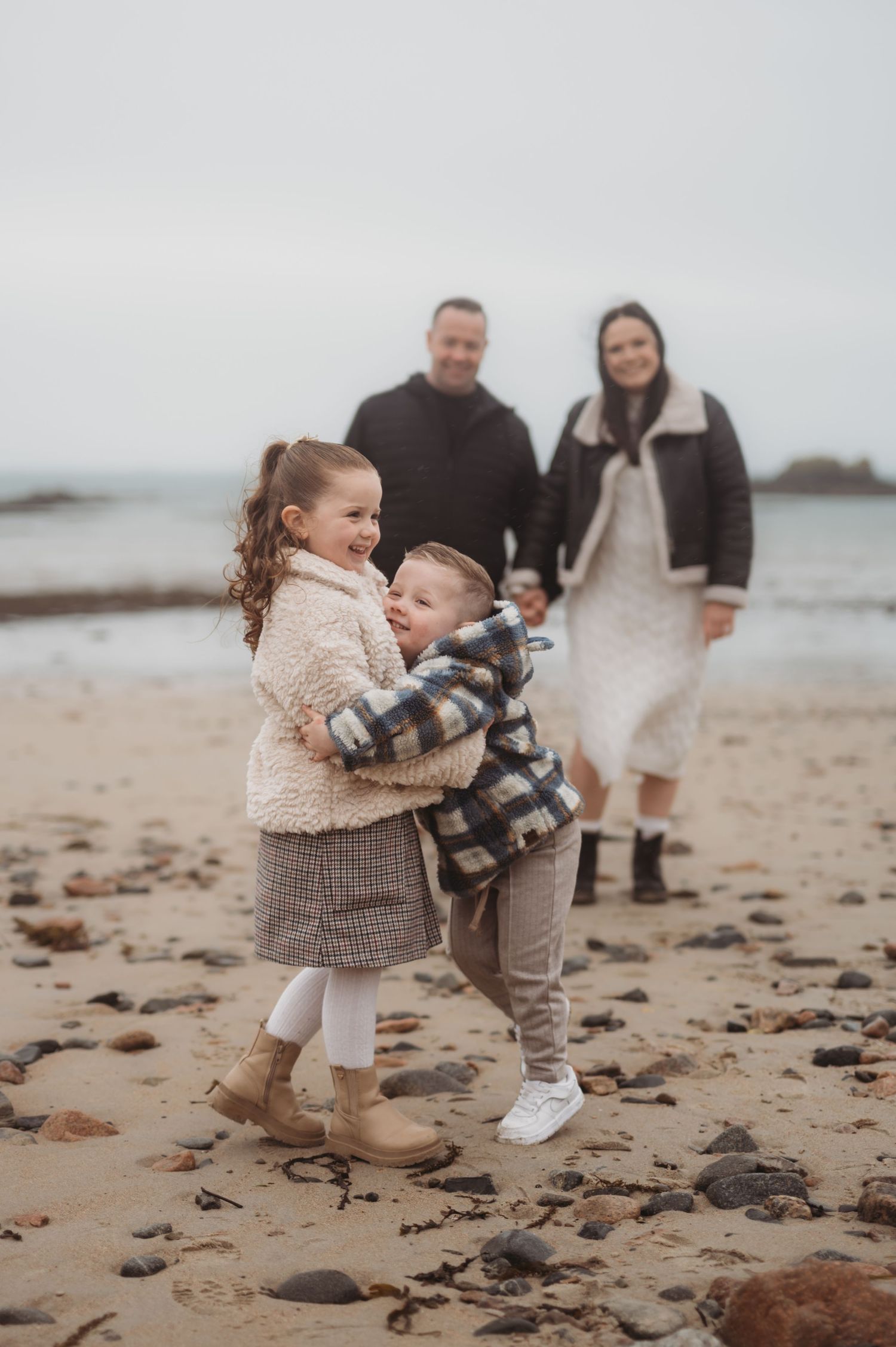 Two young siblings hugging on a rocky beach while their parents smile in the background.