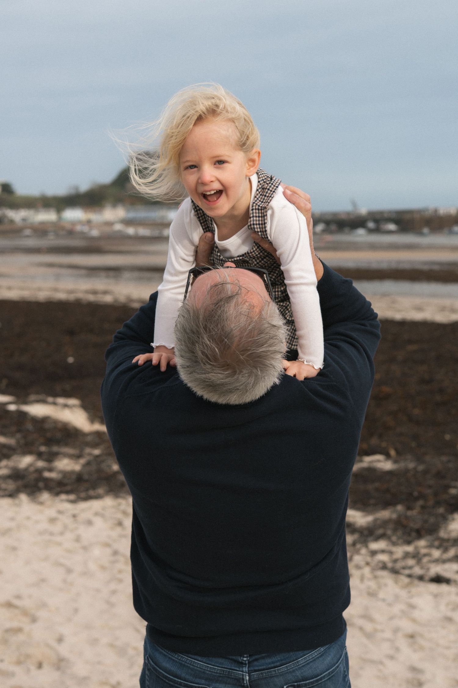 Laughing blonde toddler girl sits on her grandfather's shoulders at a beach on a cloudy day.