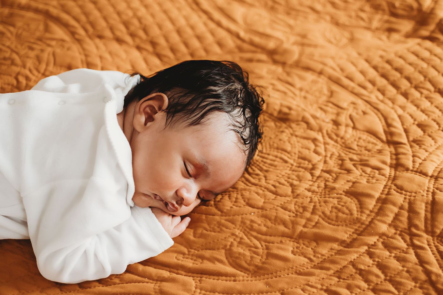 Newborn baby with dark hair sleeping peacefully in white outfit on a textured orange quilted blanket.