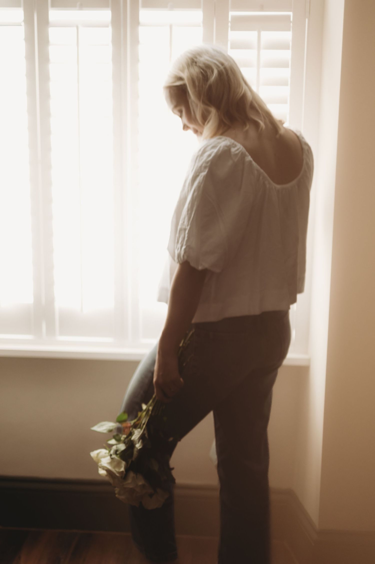 Blonde woman in white top holding flowers while standing by a bright window in soft natural light.
