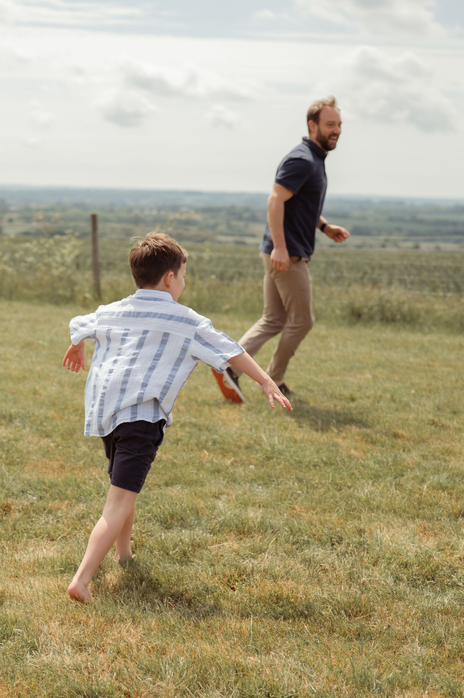 Young barefoot boy running toward his father across a grassy hillside on a sunny day.