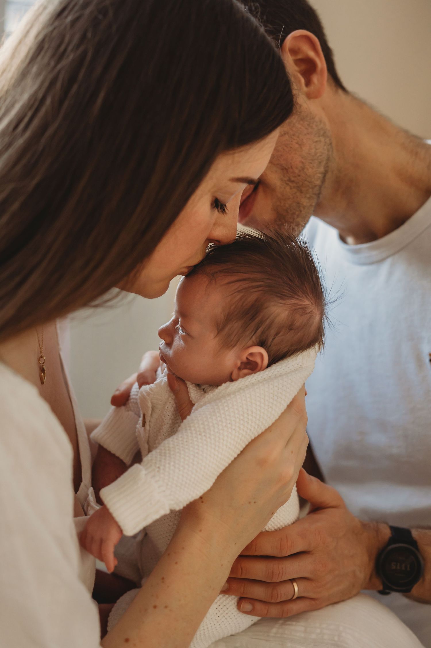 Two parents lovingly kiss their newborn baby wrapped in a white knit blanket.
