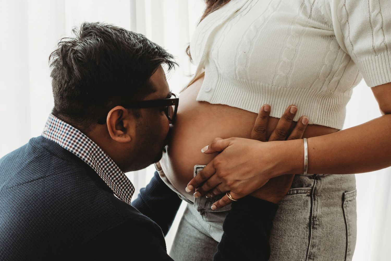 Man kissing pregnant woman's bare baby bump during intimate indoor maternity photography session.