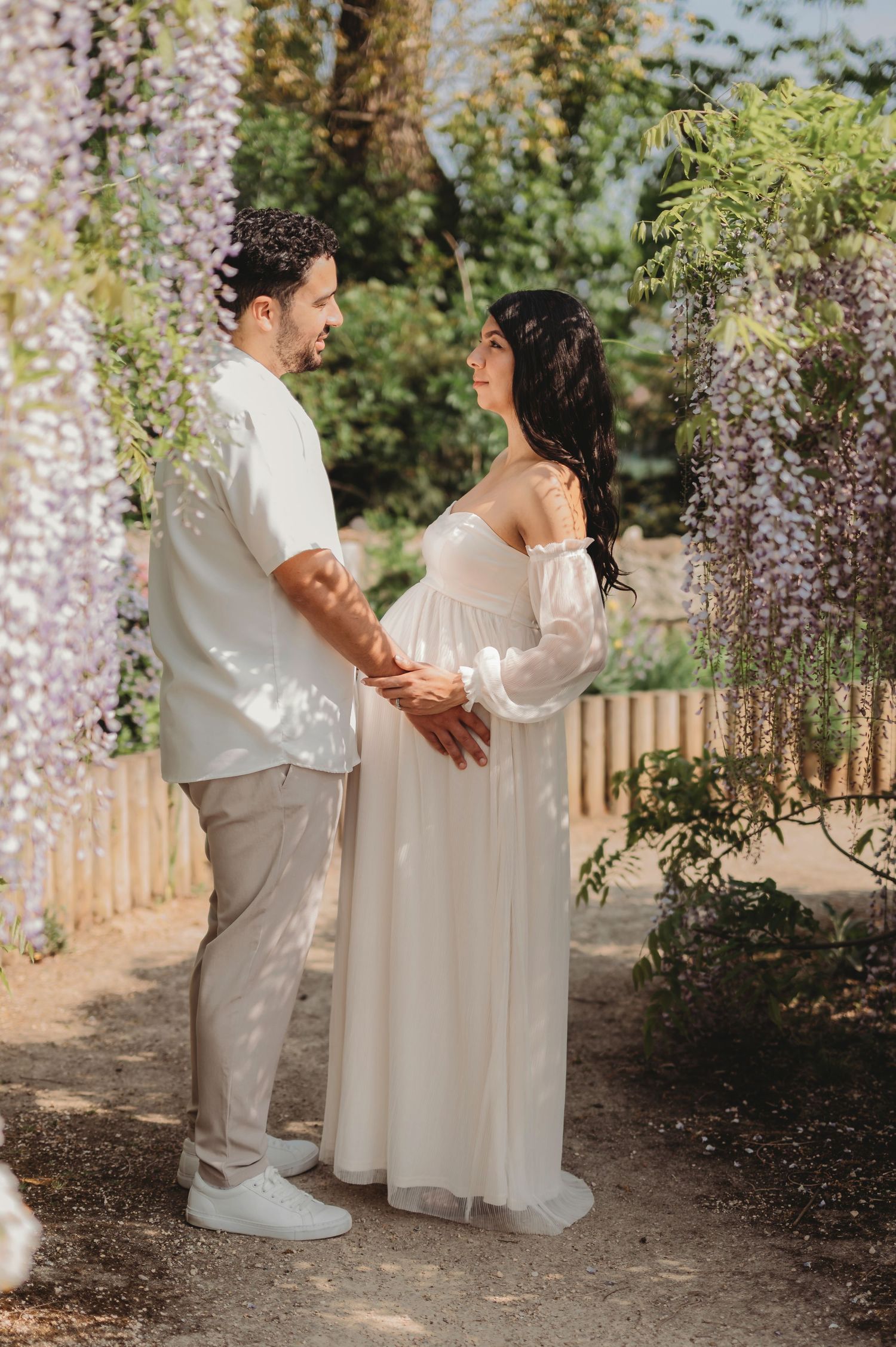 Expecting couple holds hands lovingly beneath blooming wisteria in a sunlit garden setting.