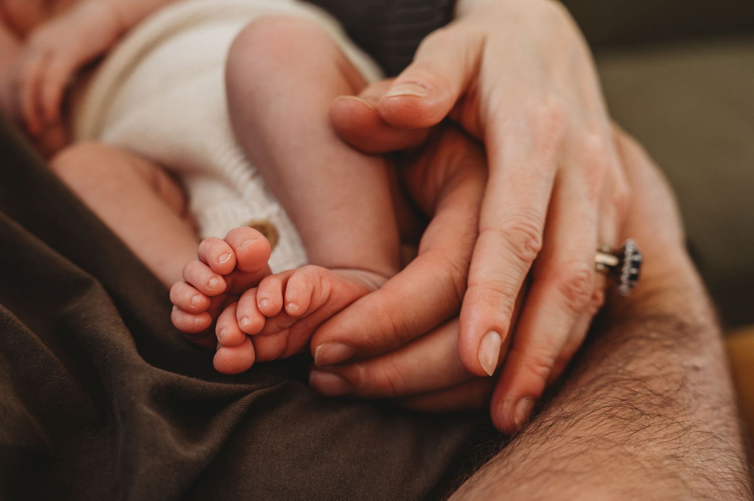 Close-up of a newborn baby's tiny feet cradled gently in parents' hands with a ring visible.