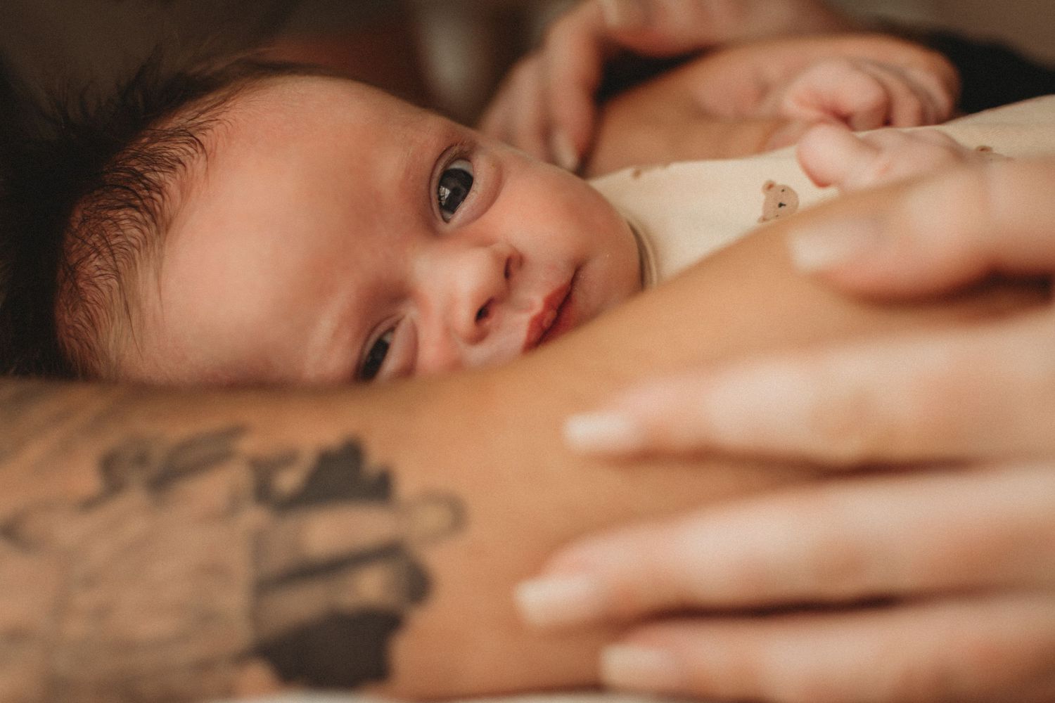 Smiling newborn baby resting on parent's arm with visible tattoo in foreground.