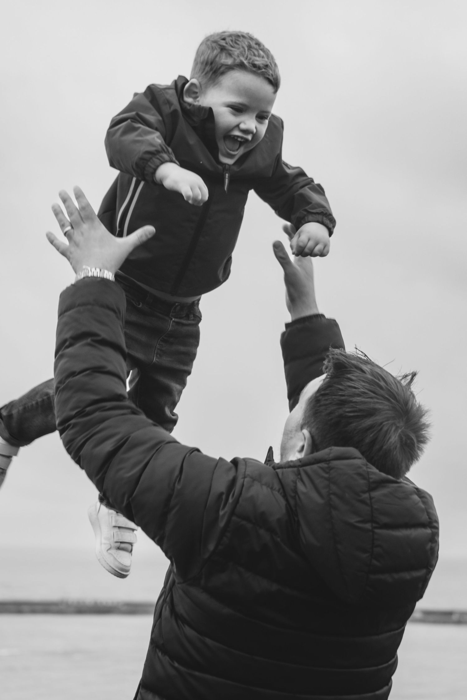 Black and white photo of a father tossing his laughing young son into the air outdoors.
