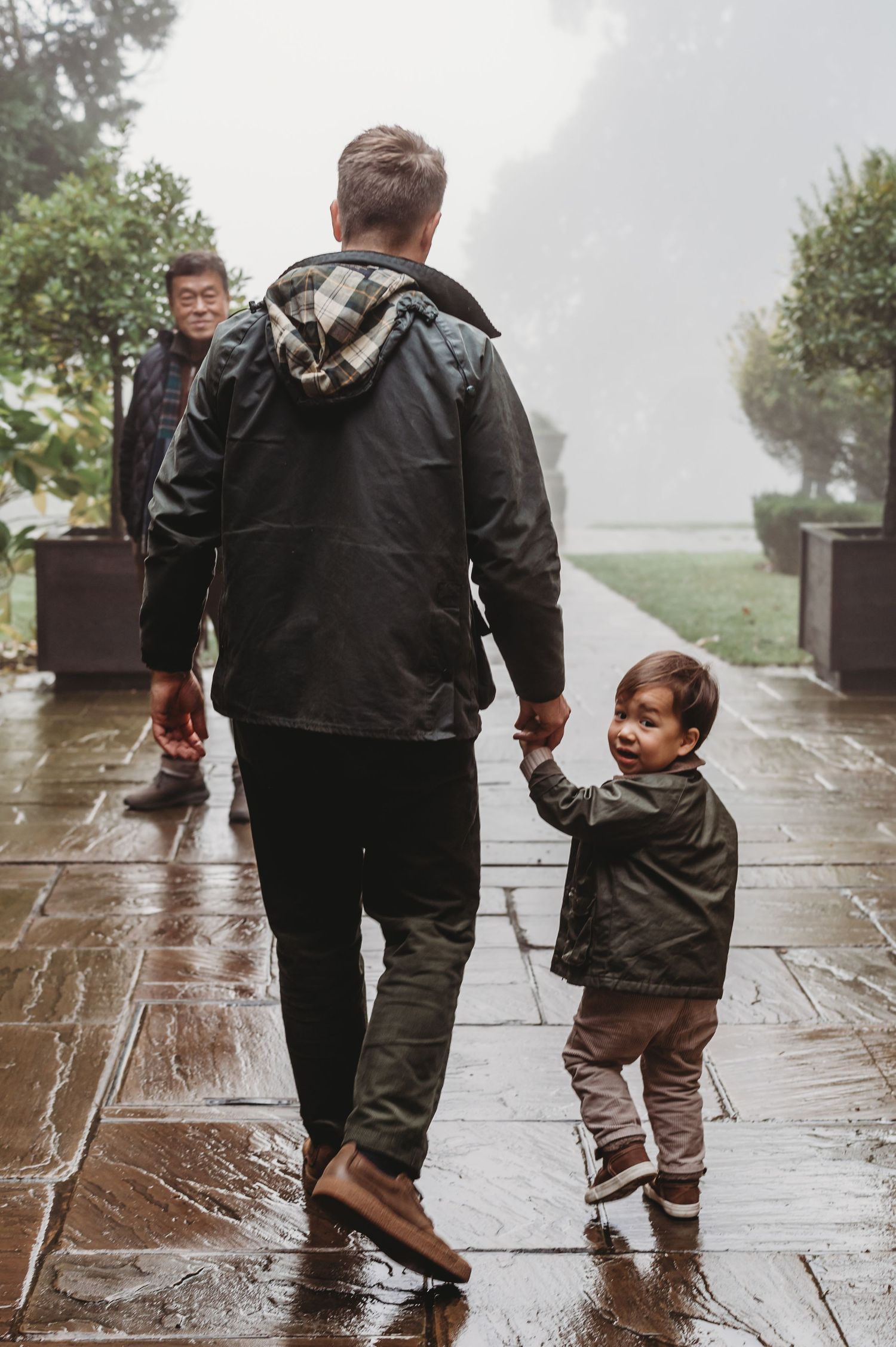 A father walks hand in hand with his smiling toddler on a rainy wet outdoor pathway.