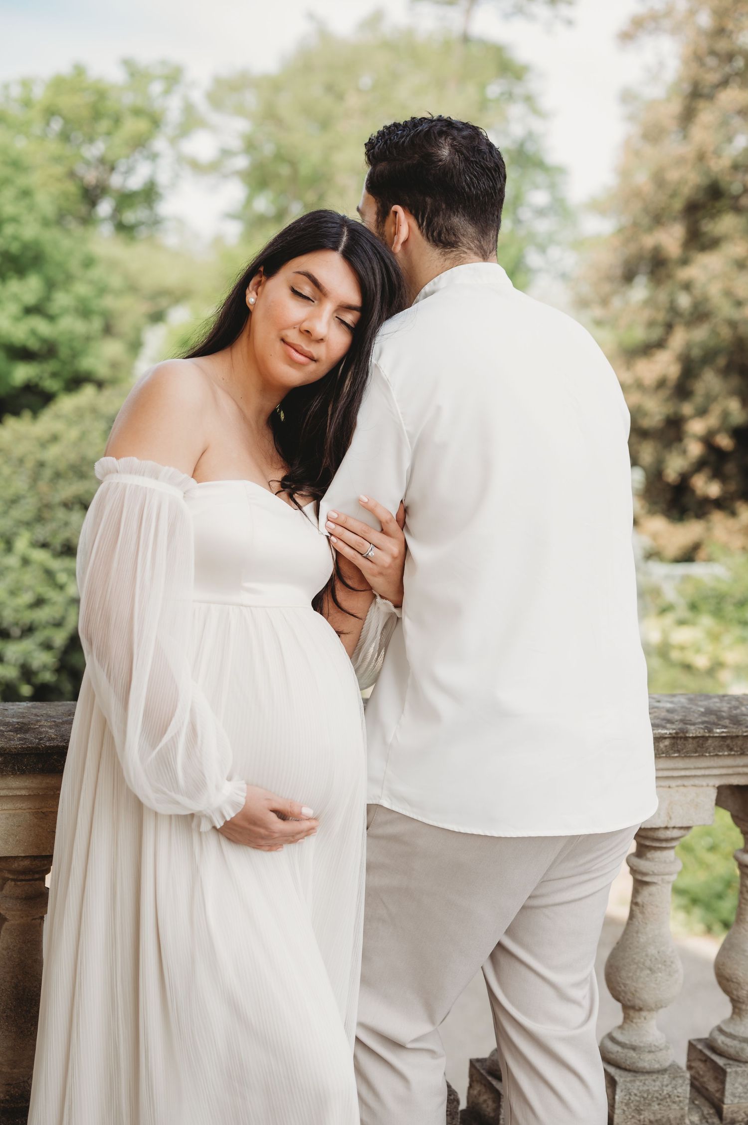 Pregnant woman in white off-shoulder gown embraced by partner in white outfit on stone balcony surrounded by greenery.
