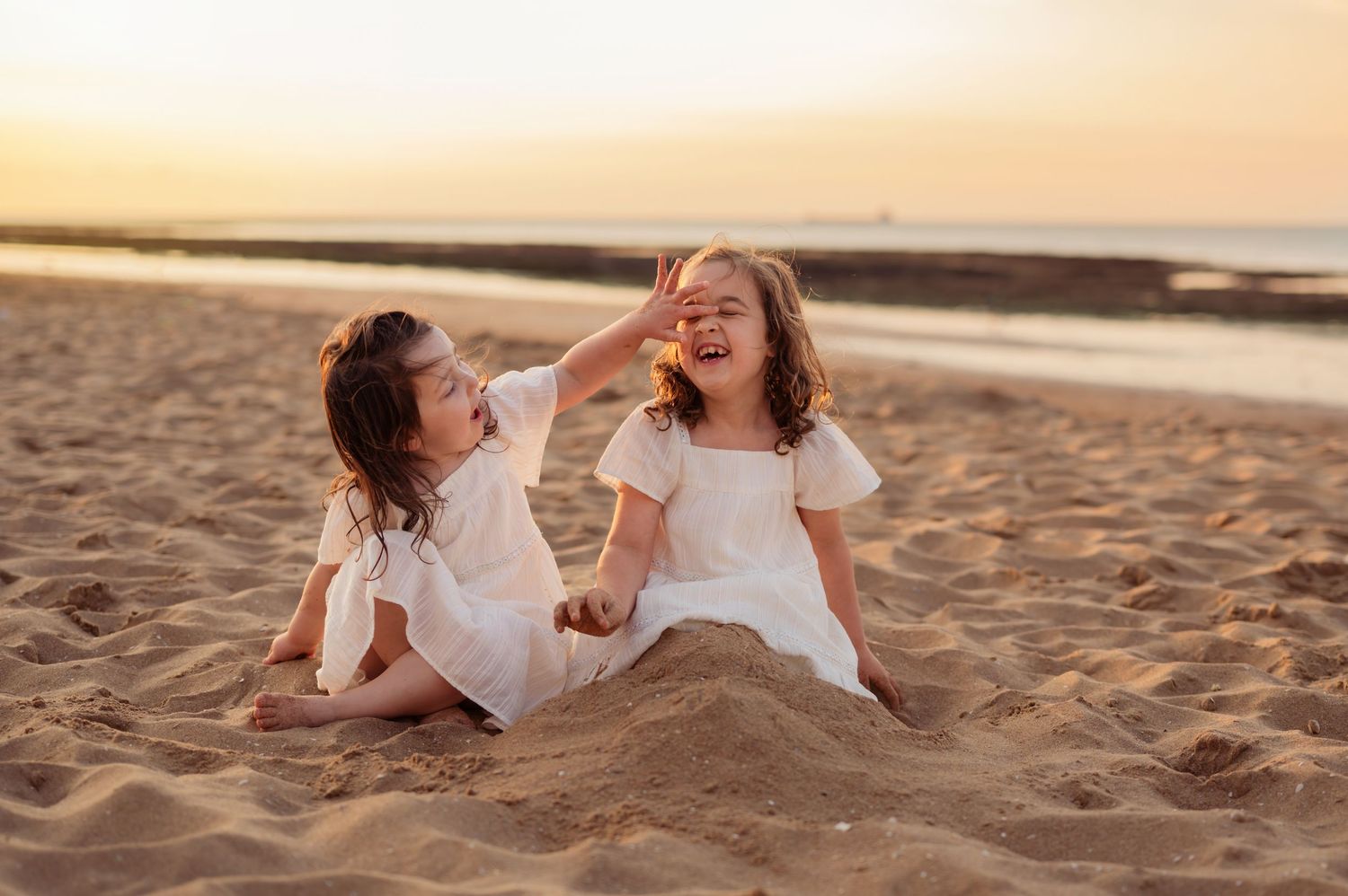 Two young sisters in white dresses laughing and playing in sand at sunset beach.
