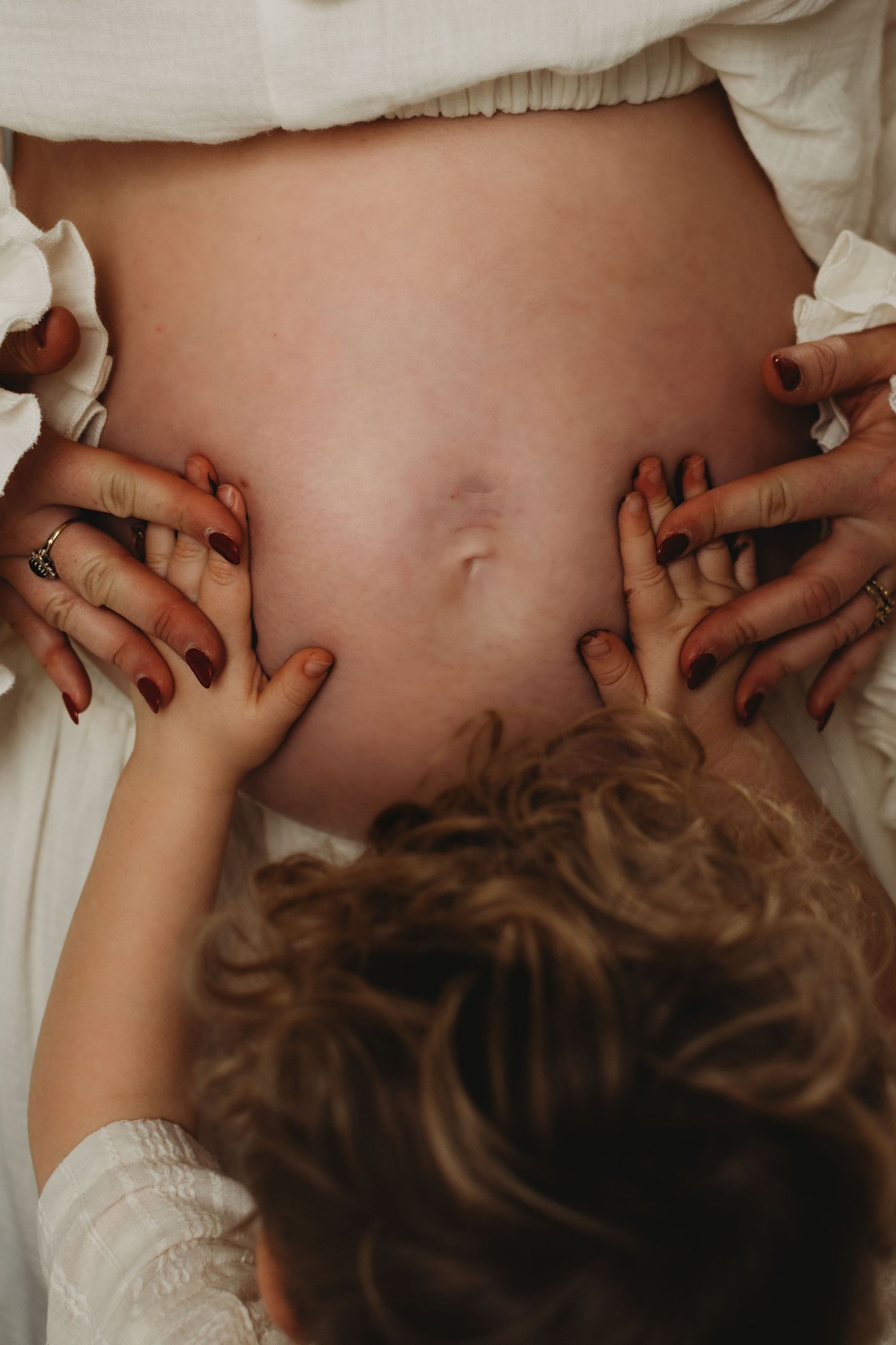 Child and mother touching pregnant belly with red-painted nails, intimate family maternity moment.