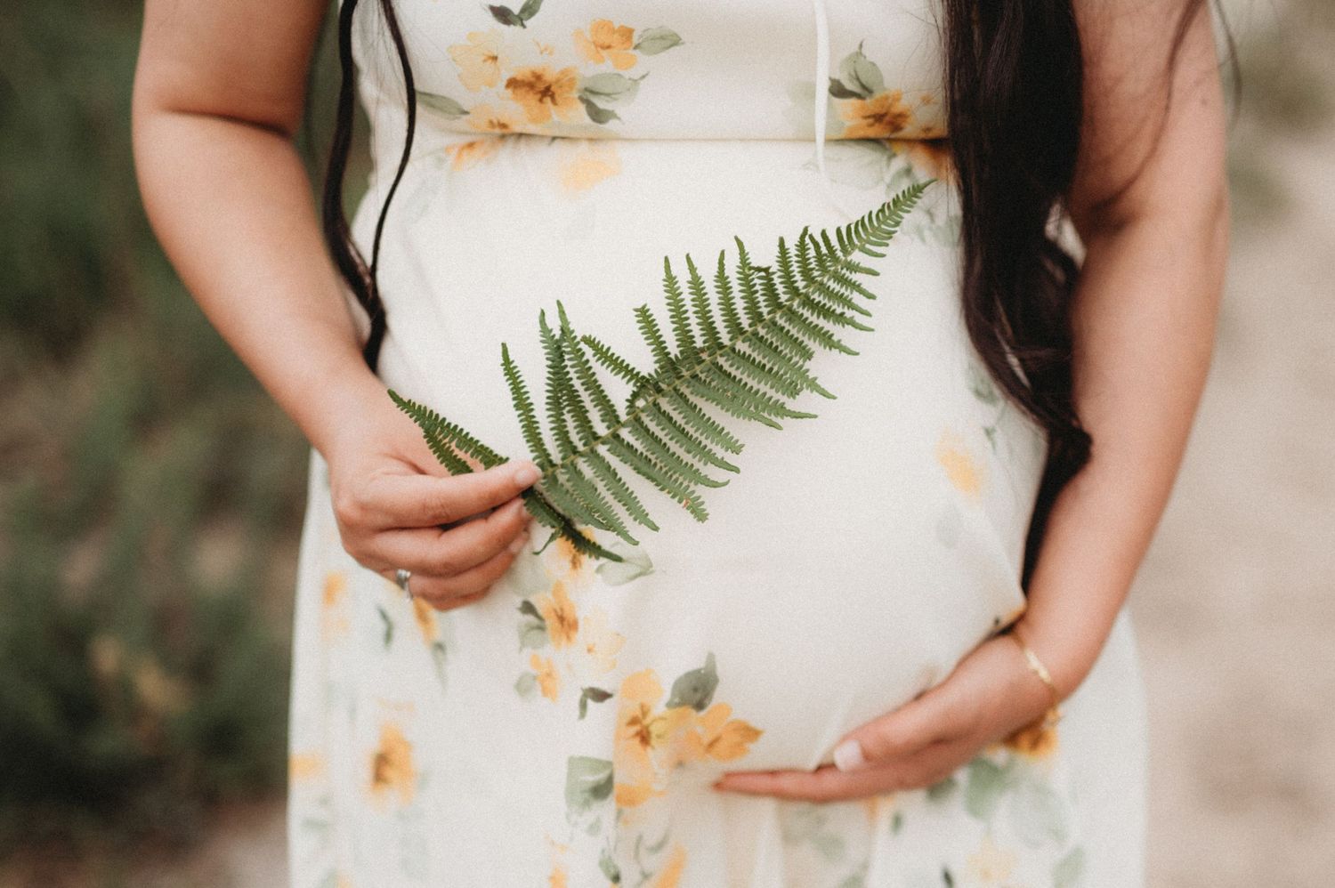 Close-up of pregnant belly in floral dress with green fern leaf.