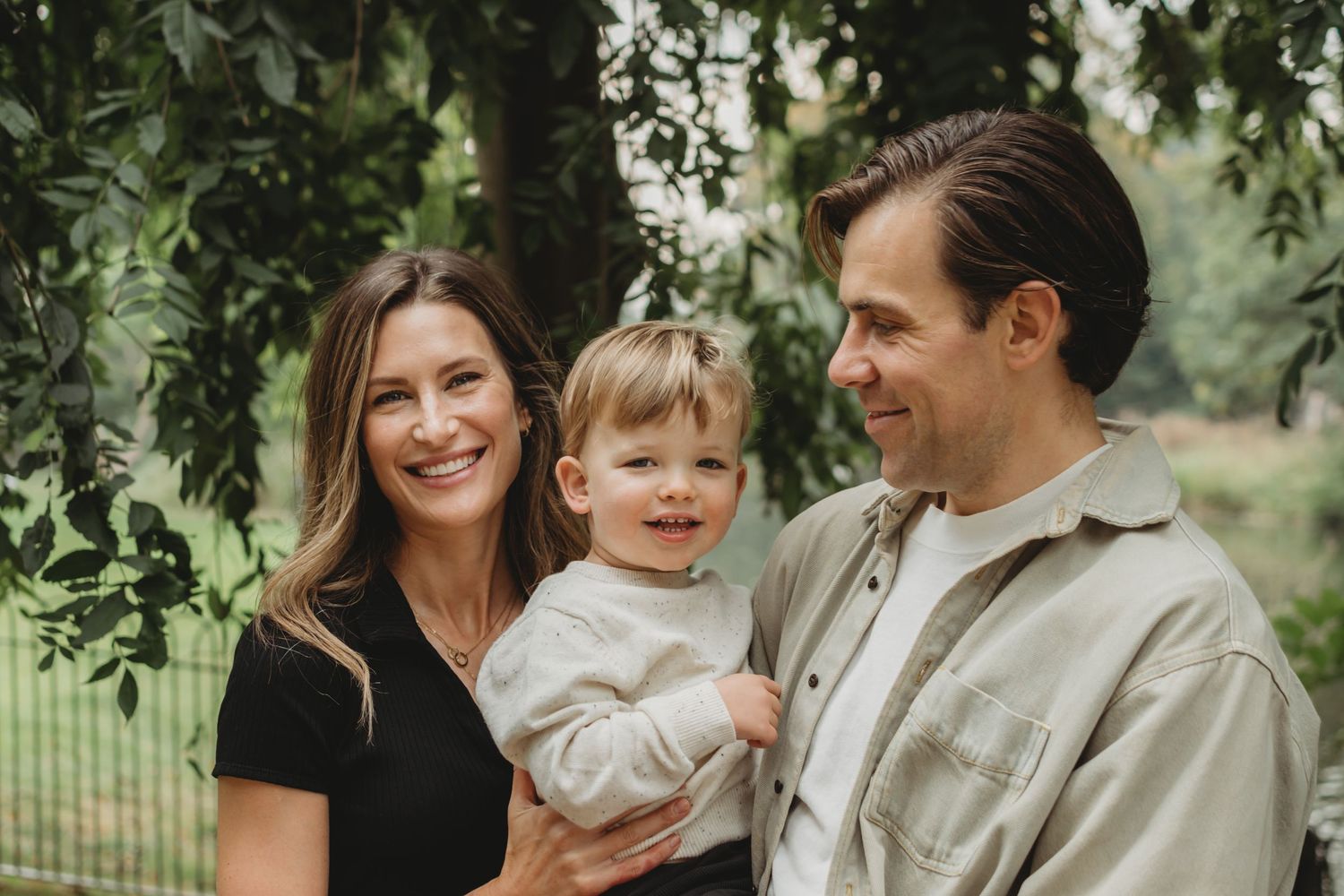 Happy young family with toddler son smiling together outdoors surrounded by lush green trees.