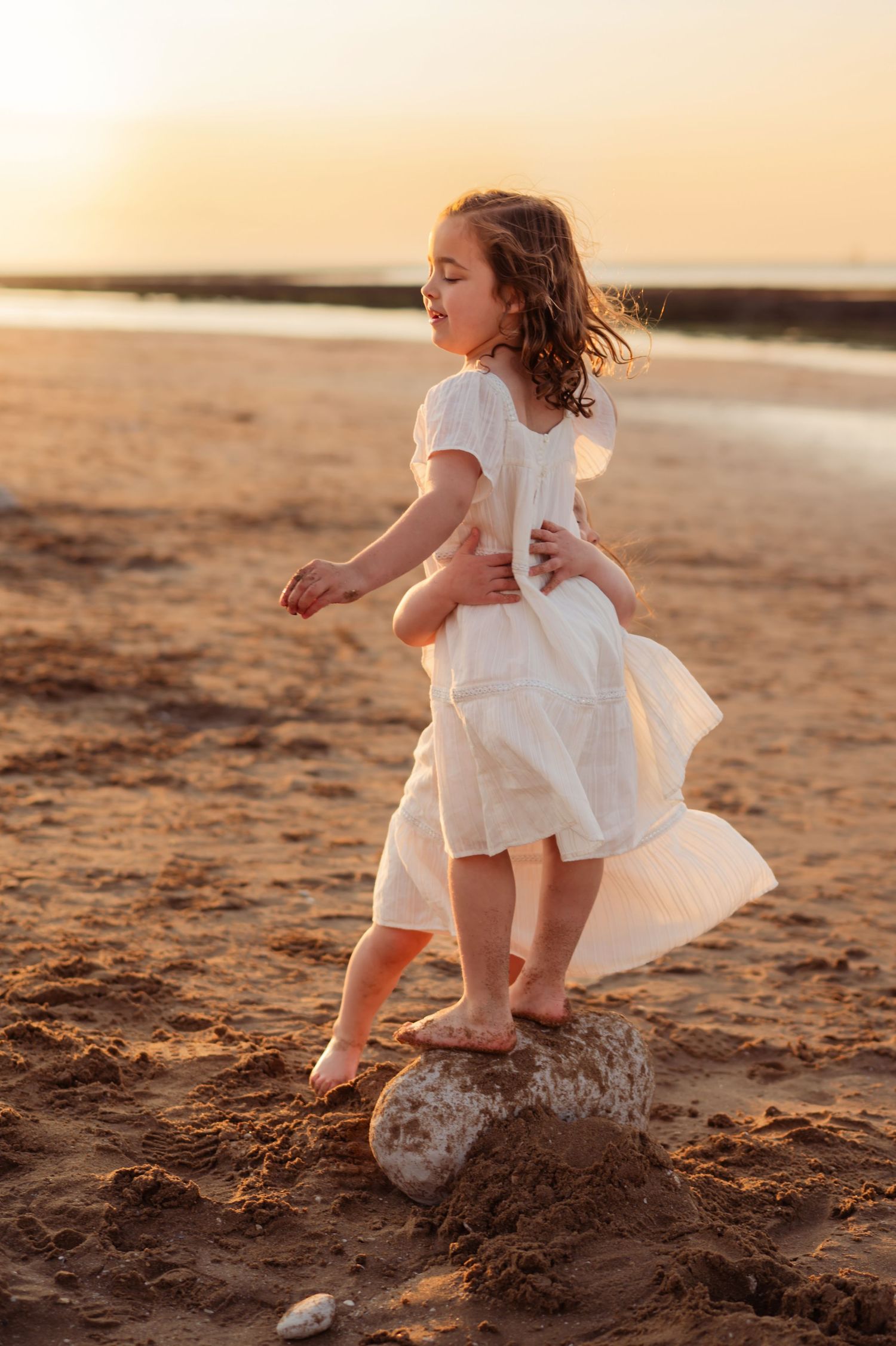 Young girl in flowing white dress standing on a rock on a sandy beach at golden sunset.