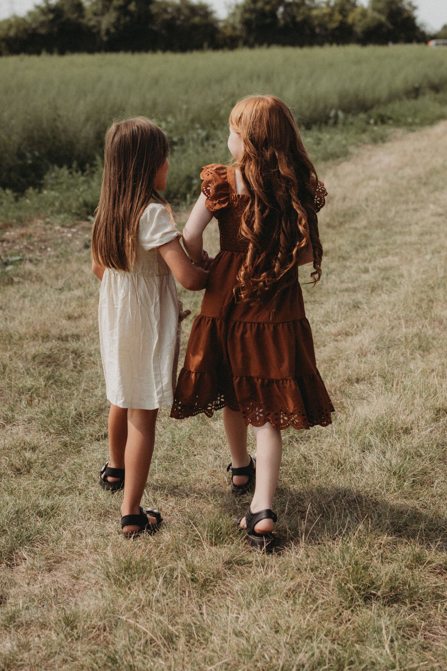 Two girls in dresses walking hand in hand along a grassy field path.