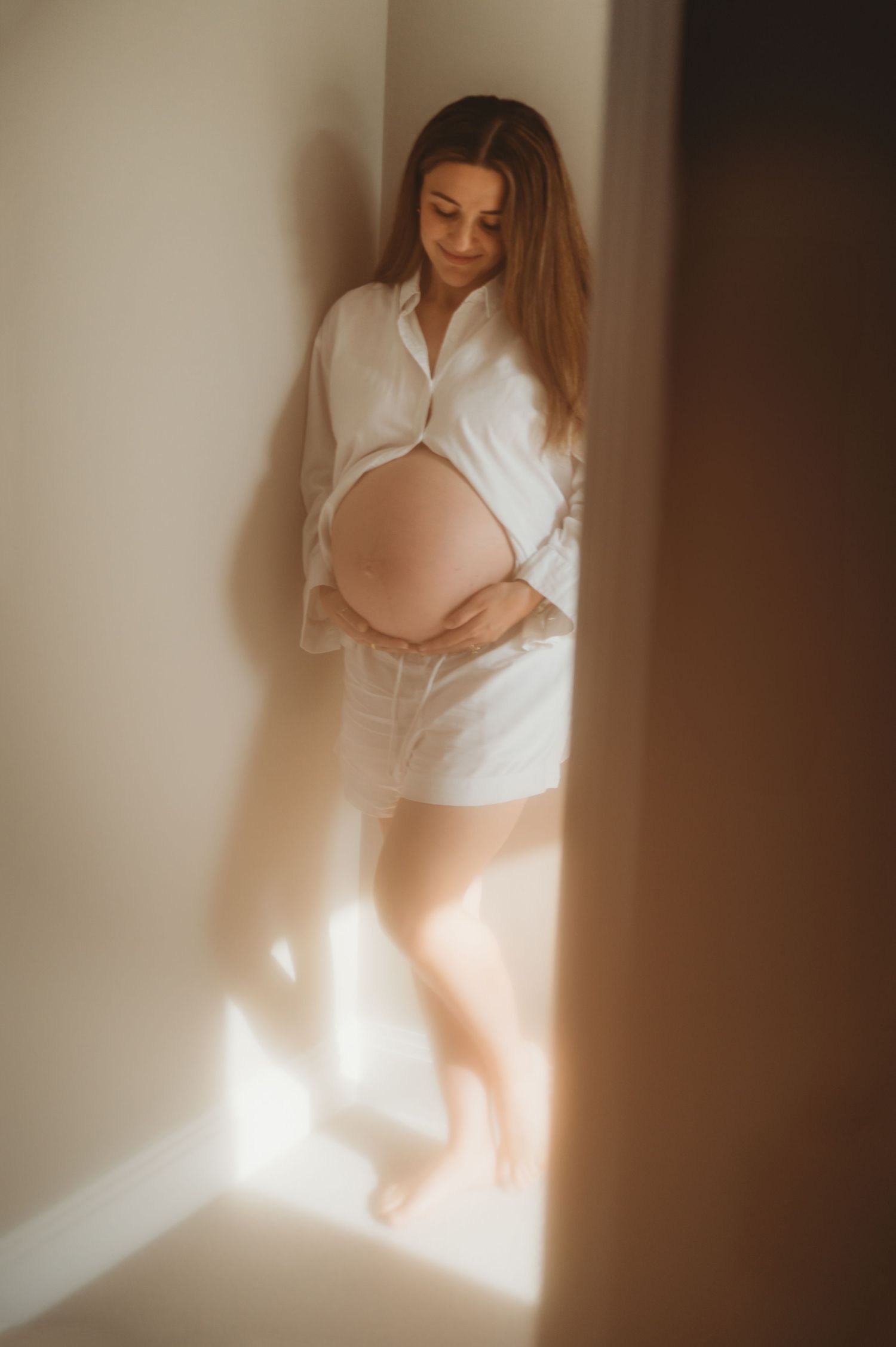 Pregnant woman in white shirt holding baby bump in warm sunlit corner.