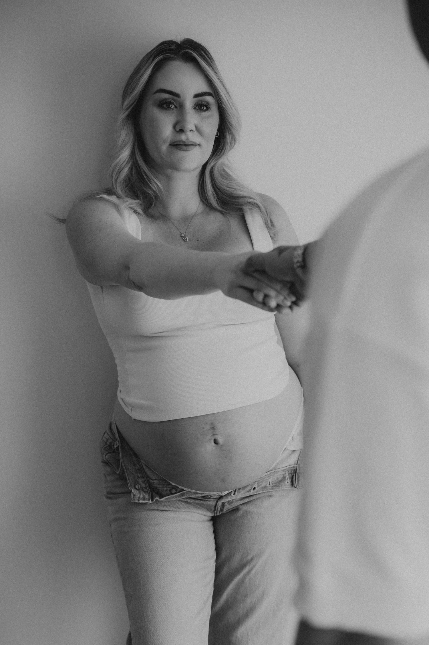Black and white photo of a pregnant blonde woman in a white tank top holding hands with her partner.