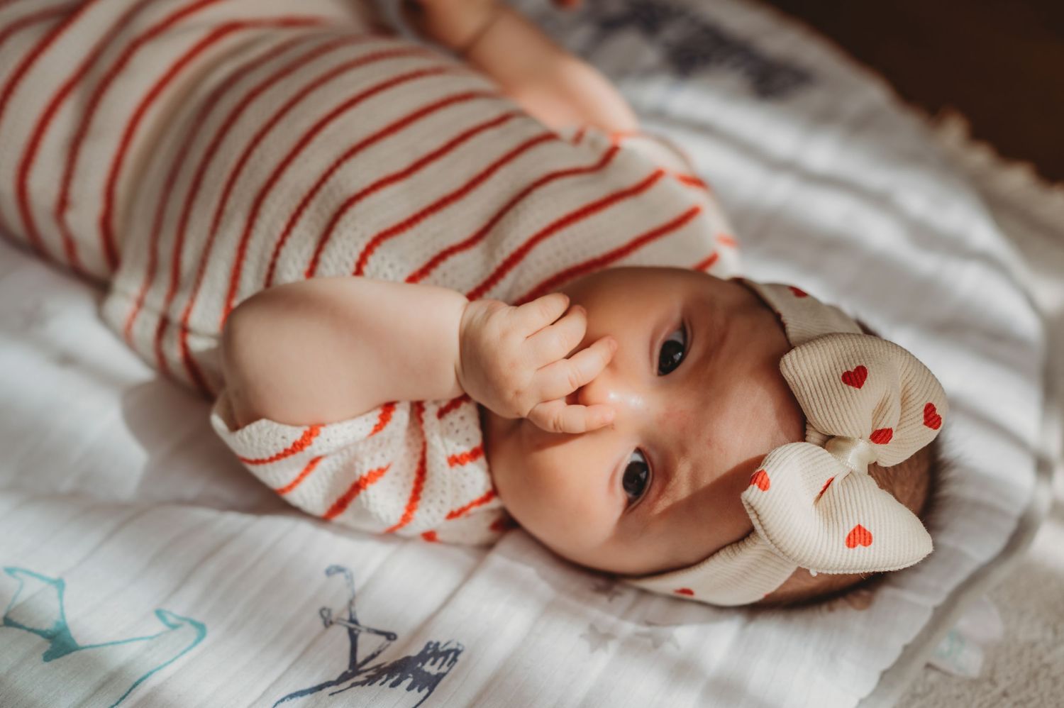 Cute baby girl in red striped onesie and heart bow headband lying on a soft blanket.