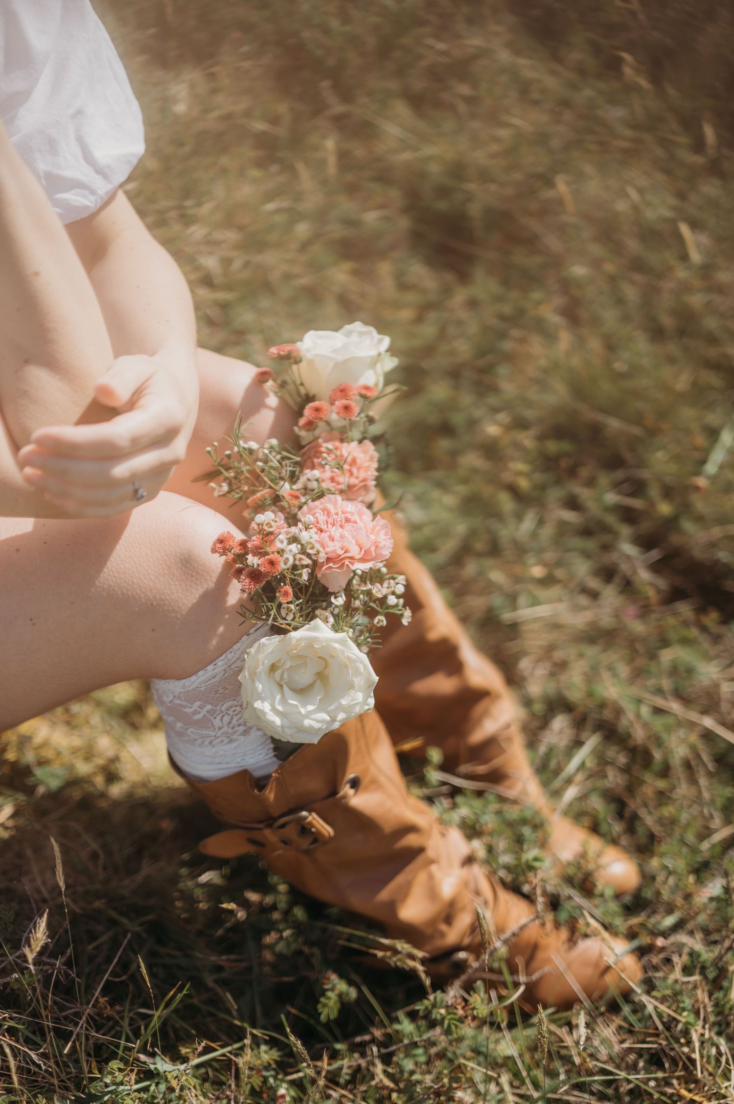 Woman sitting in grass holding delicate pink and white floral bouquet wearing brown leather boots.