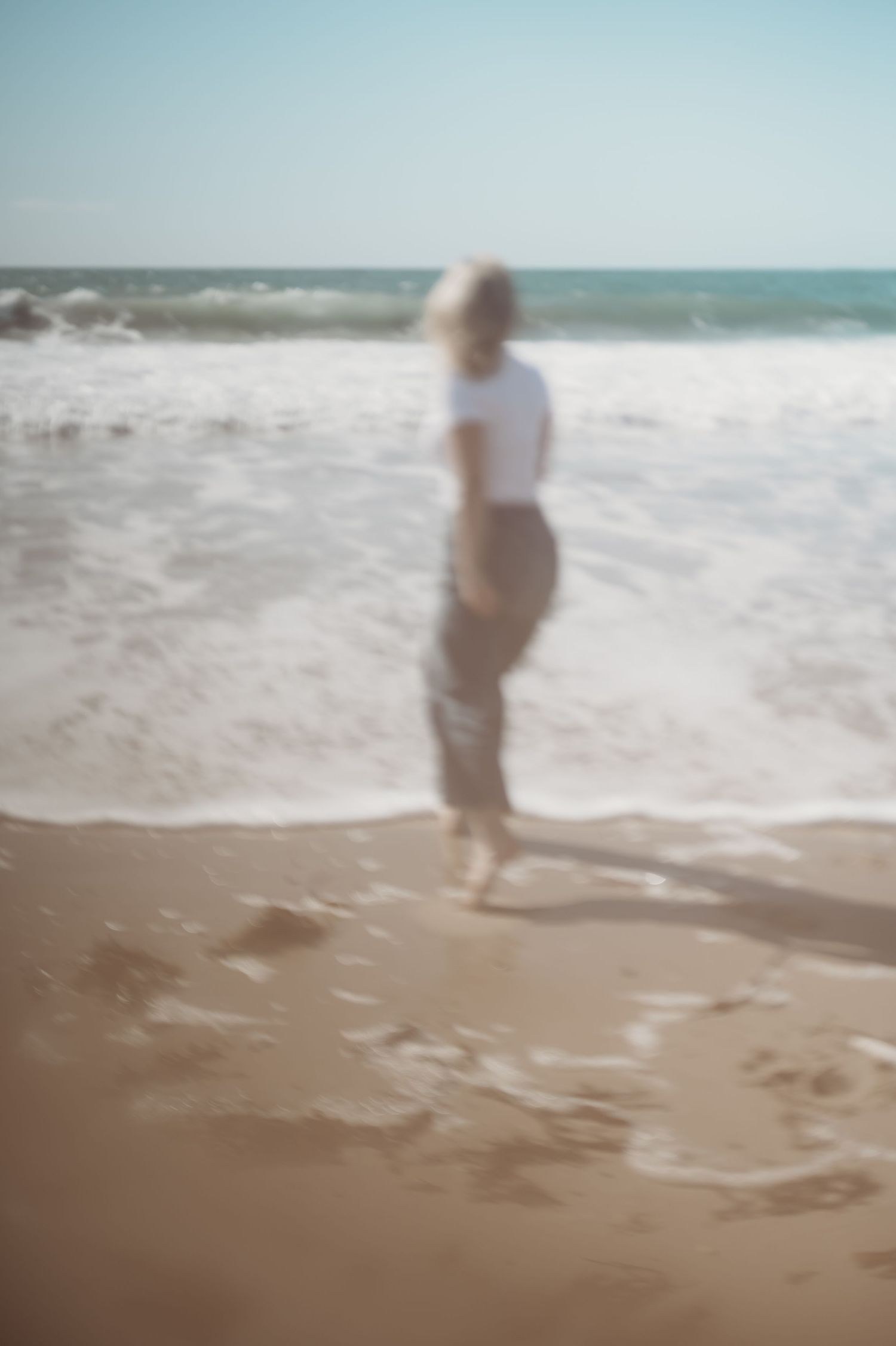 Blurred motion photo of blonde woman in white top walking toward ocean waves on beach.
