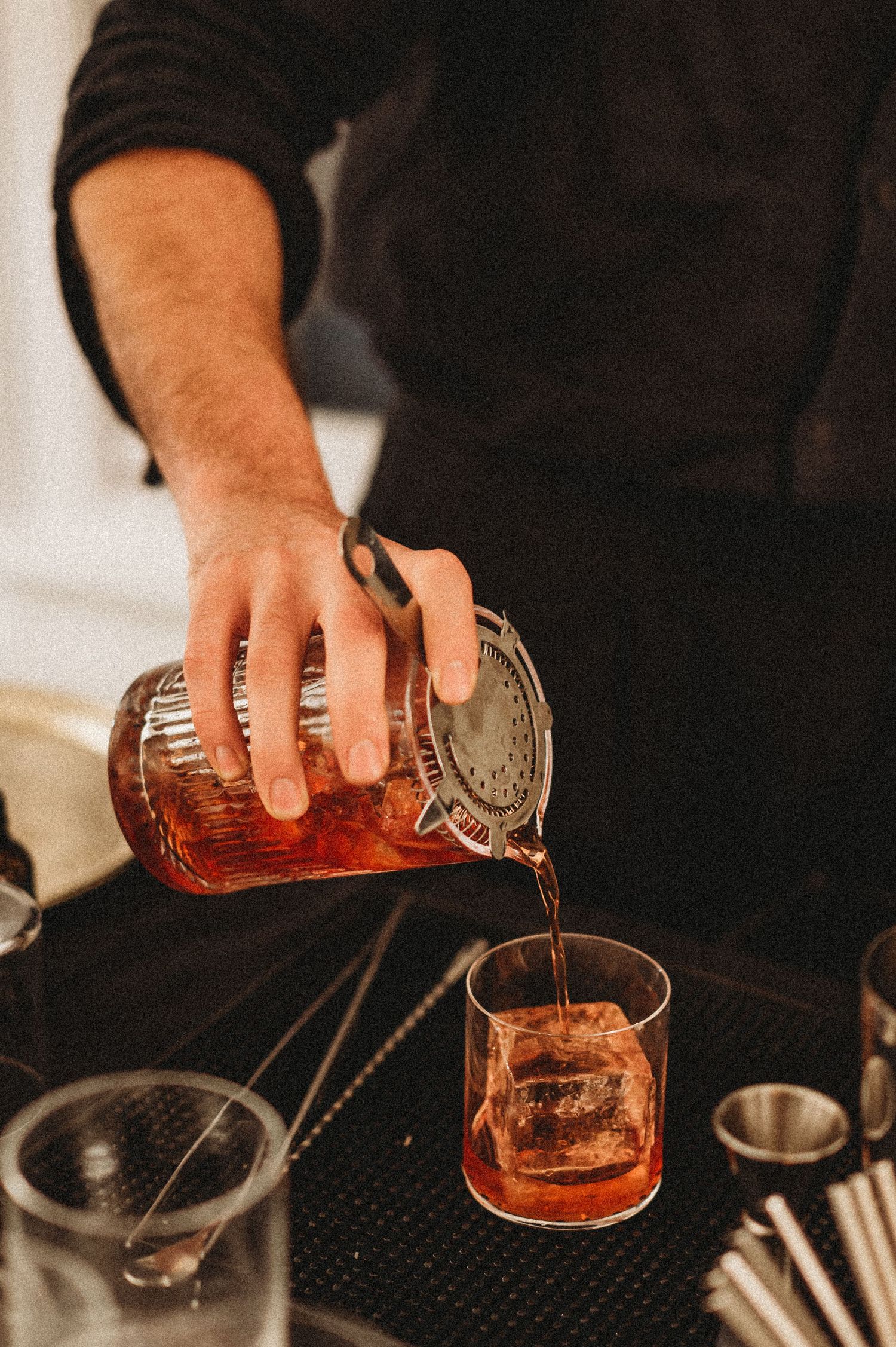 Bartender pouring a rich amber cocktail through a strainer into a rocks glass with ice.