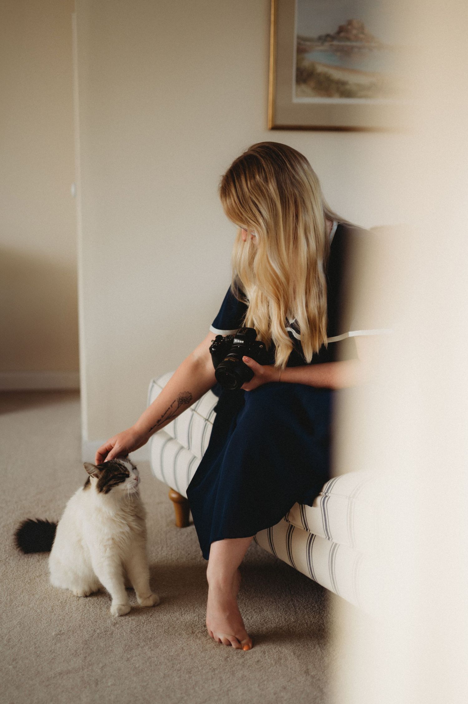 Blonde woman in navy dress holding camera while petting a fluffy cat beside a white armchair.