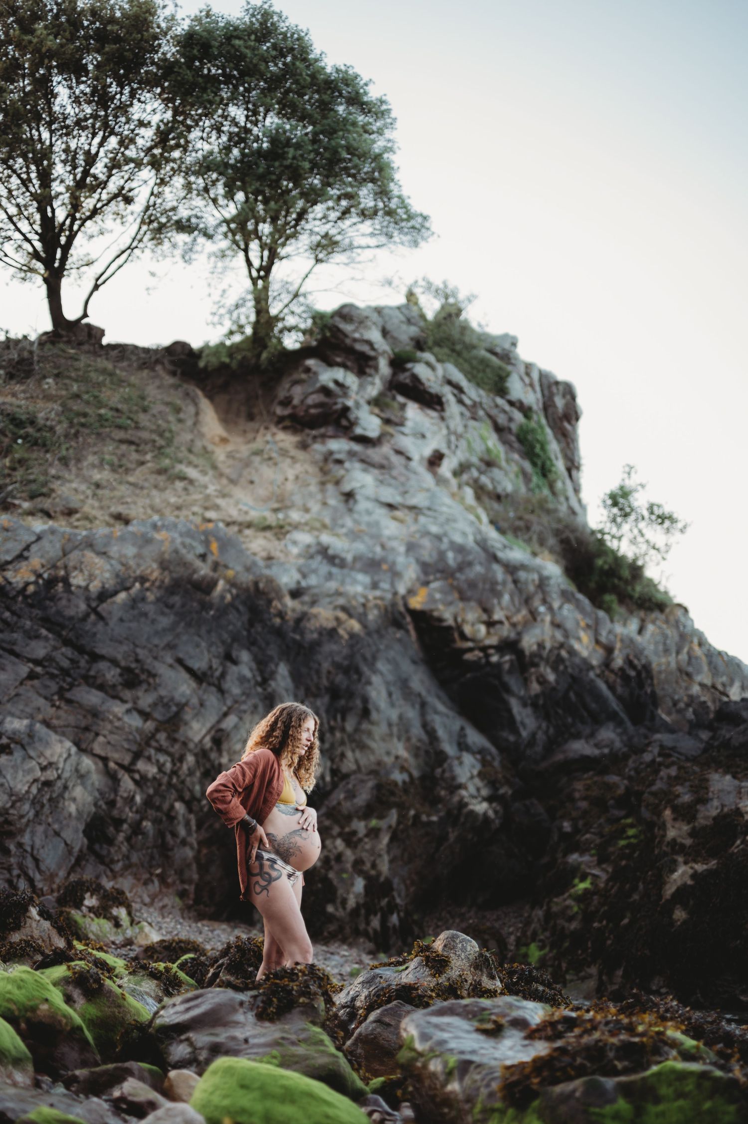 Pregnant woman in floral bikini and pink cardigan standing on rocky coastal cliffs with trees above.
