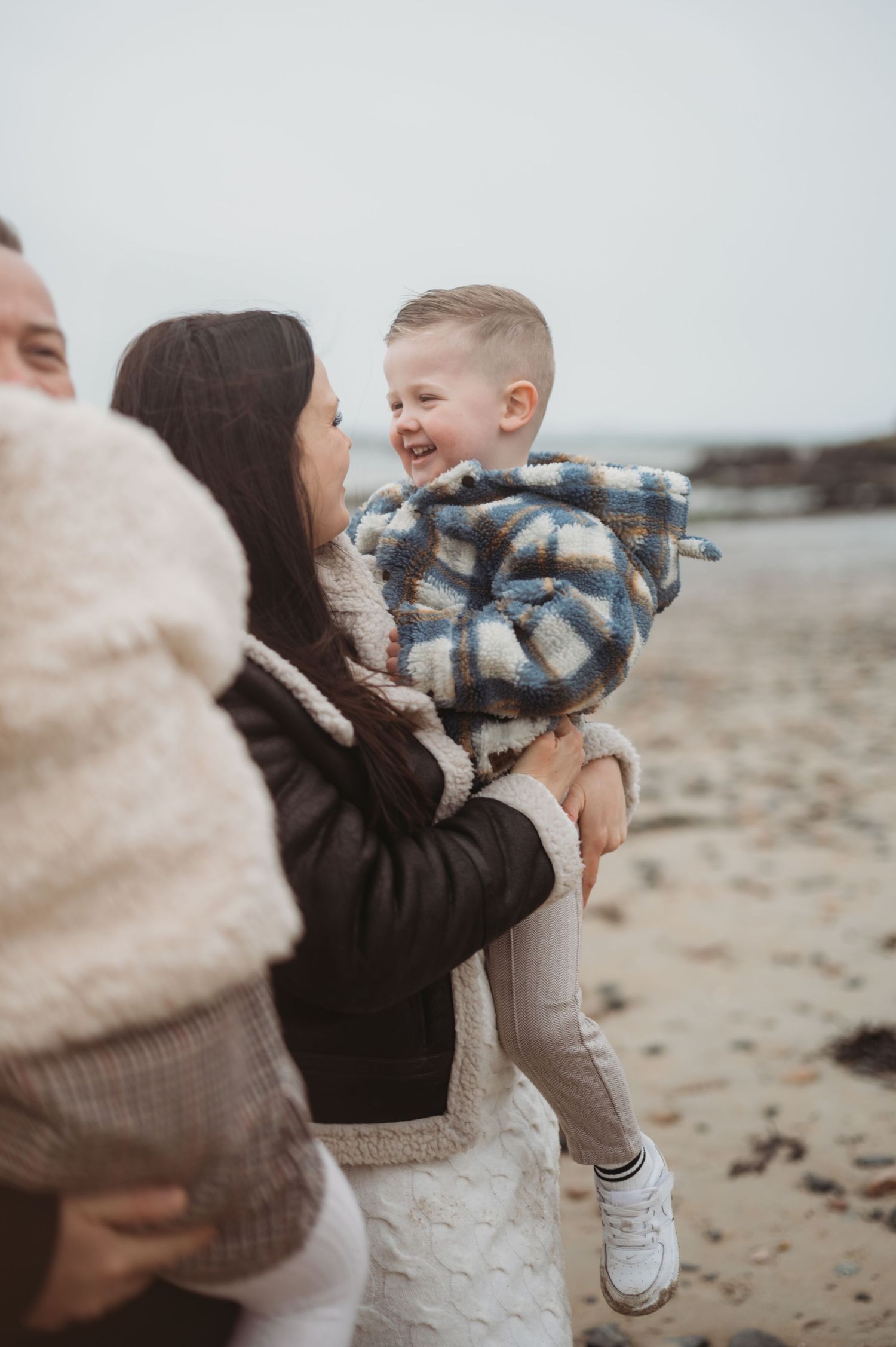 A laughing toddler in a plaid jacket is held by his mother on a cold beach while father stands nearby.