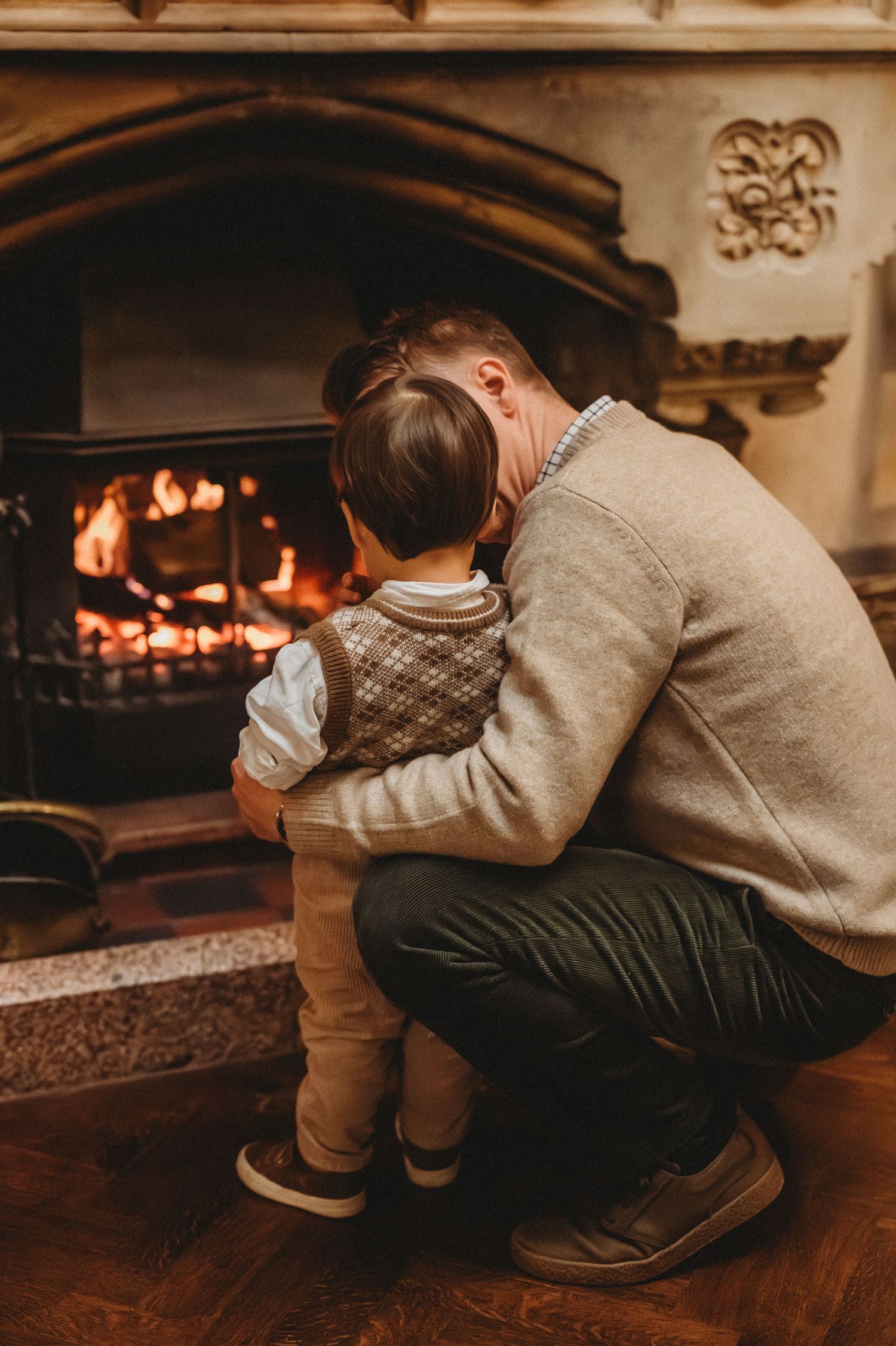 A father crouches down hugging his young child in front of a warm glowing fireplace indoors.