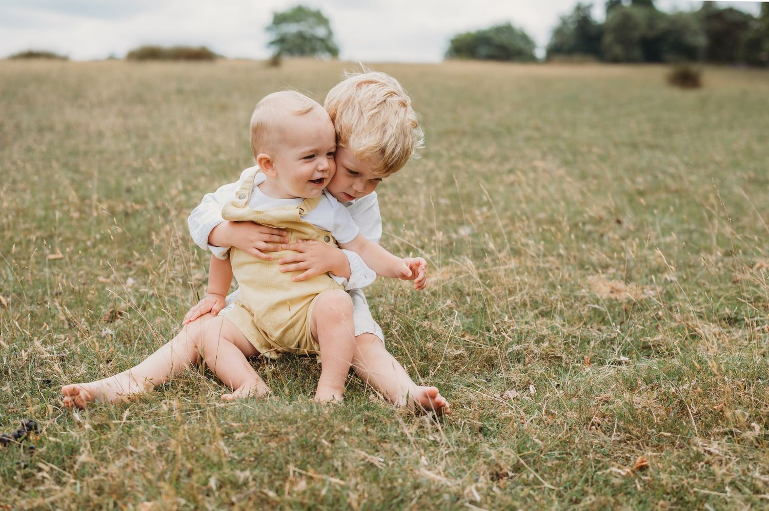 Two young siblings sit together in a grassy field, the older child hugging the baby warmly.