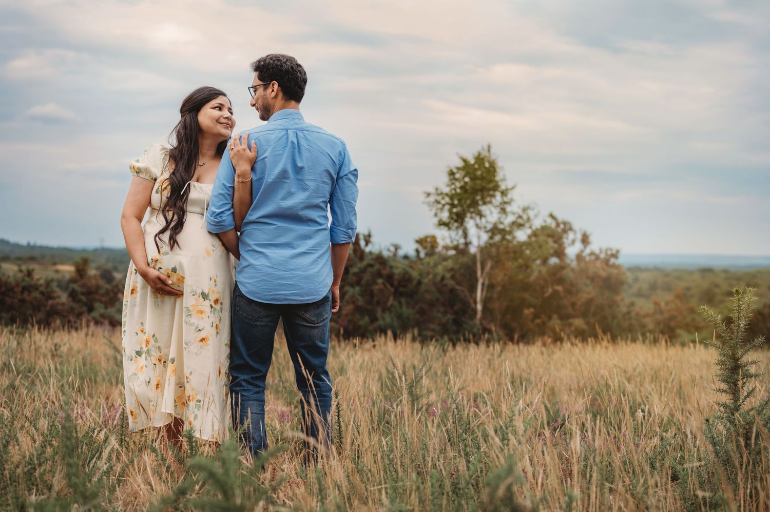 Pregnant woman and partner standing in tall grass field, sharing a tender moment during maternity photoshoot.
