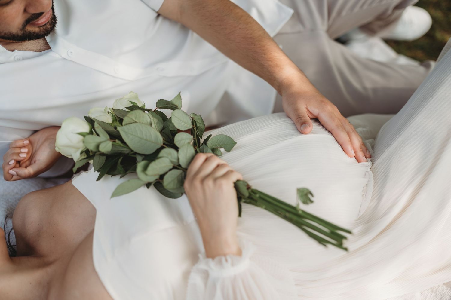 Close-up of pregnant belly in white dress with eucalyptus bouquet held by couple sitting together outdoors.