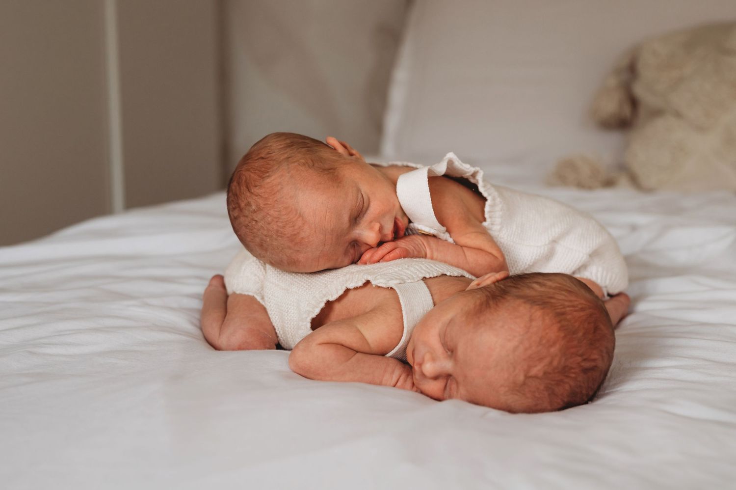 Two newborn twins in white onesies sleeping peacefully side by side on a white bed.
