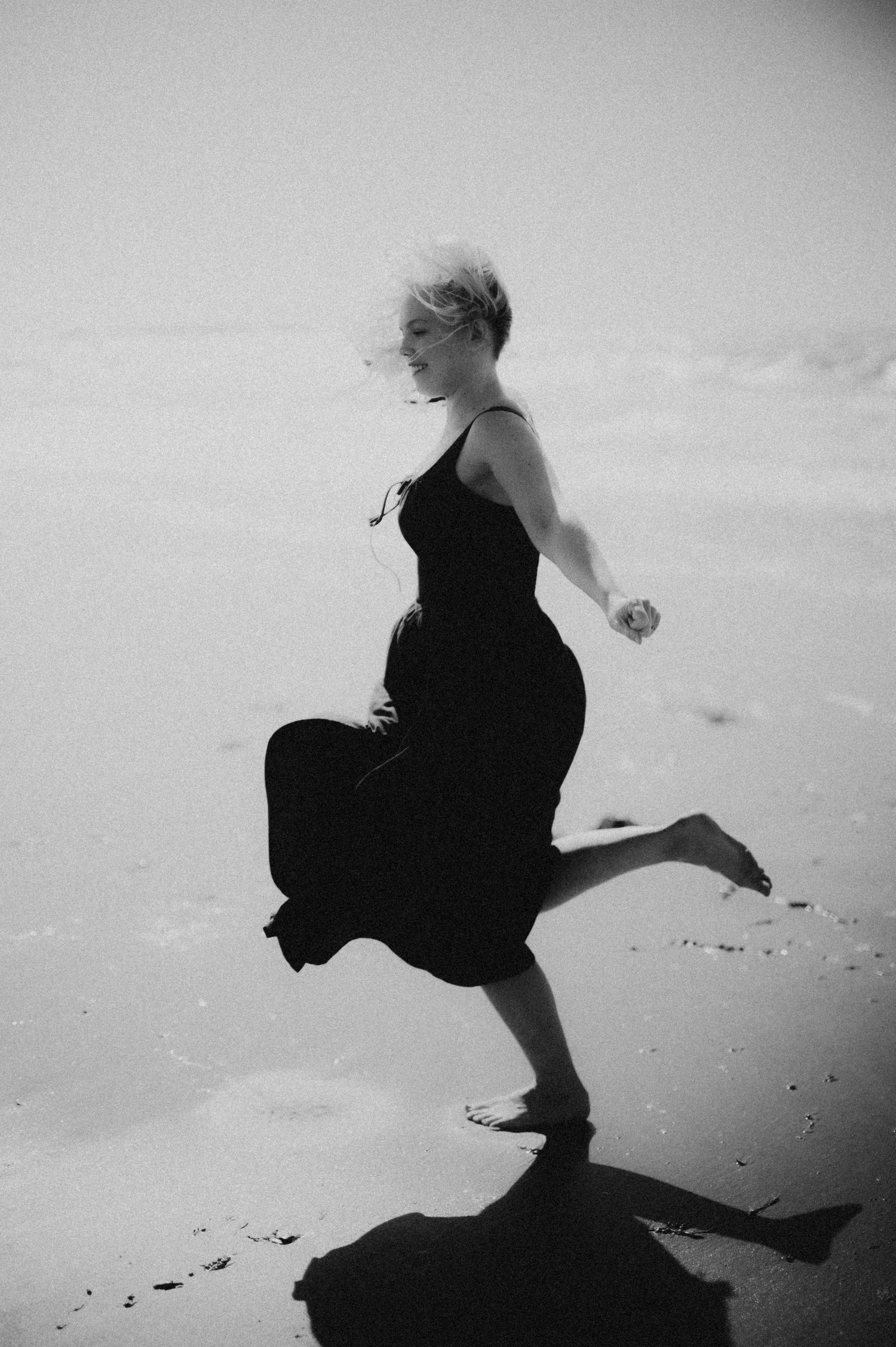 Black and white photo of blonde woman in black dress running barefoot on beach.