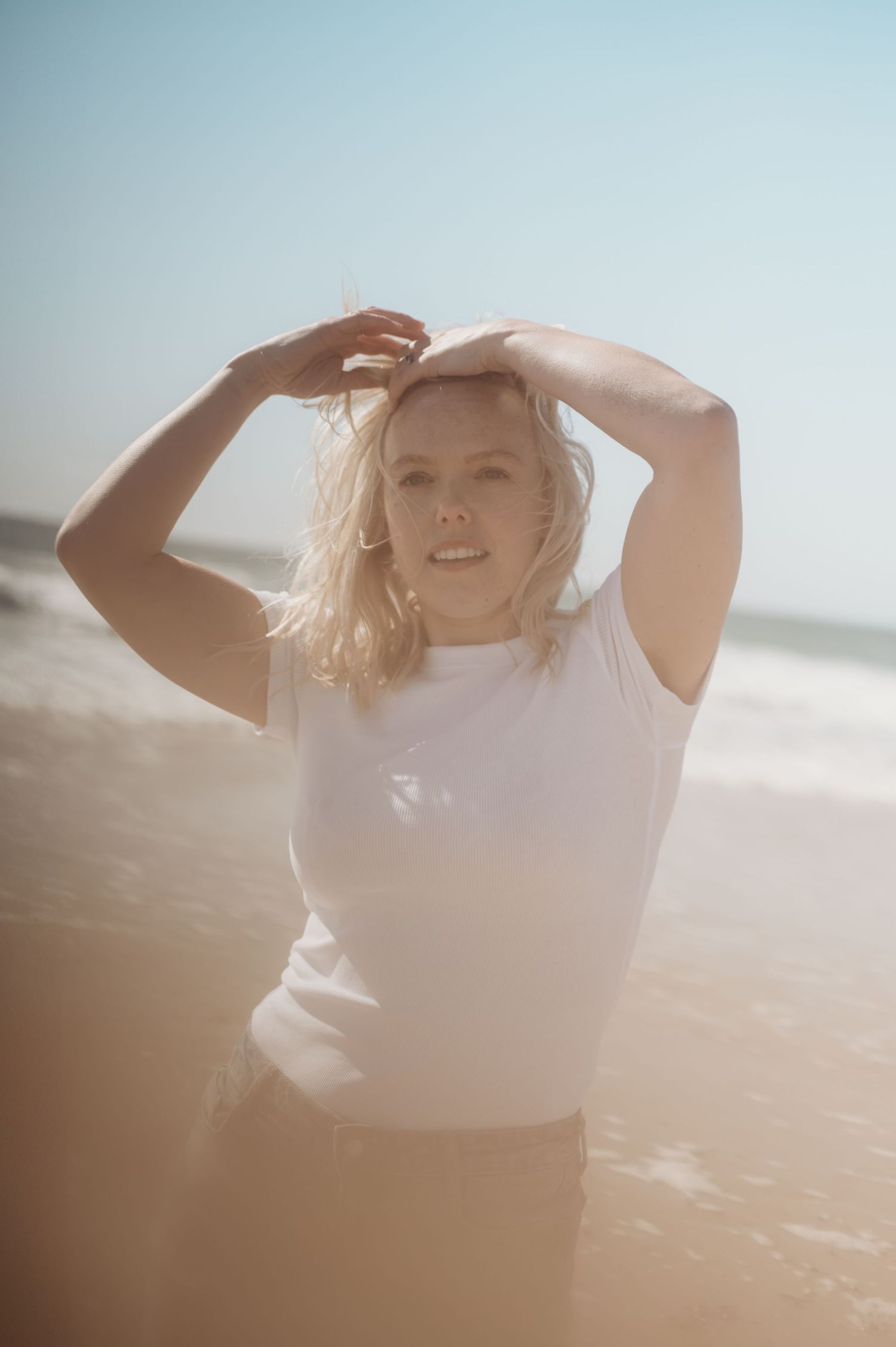 Smiling blonde woman in white sleeveless top standing in ocean waves on a sunny beach.