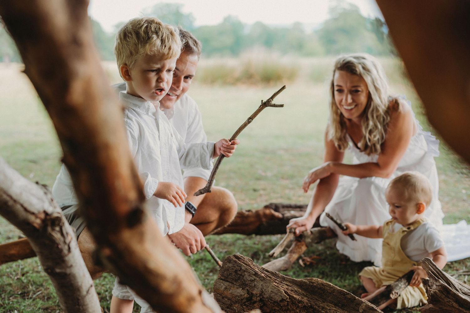 Children and woman playing with sticks near log structure in grassy outdoor area.