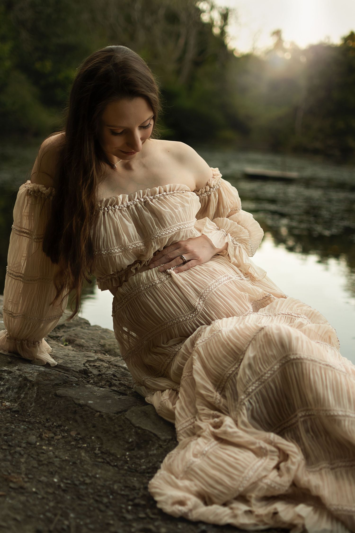 Pregnant woman in flowing boho cream dress sitting by a serene lake surrounded by lush greenery at sunset.
