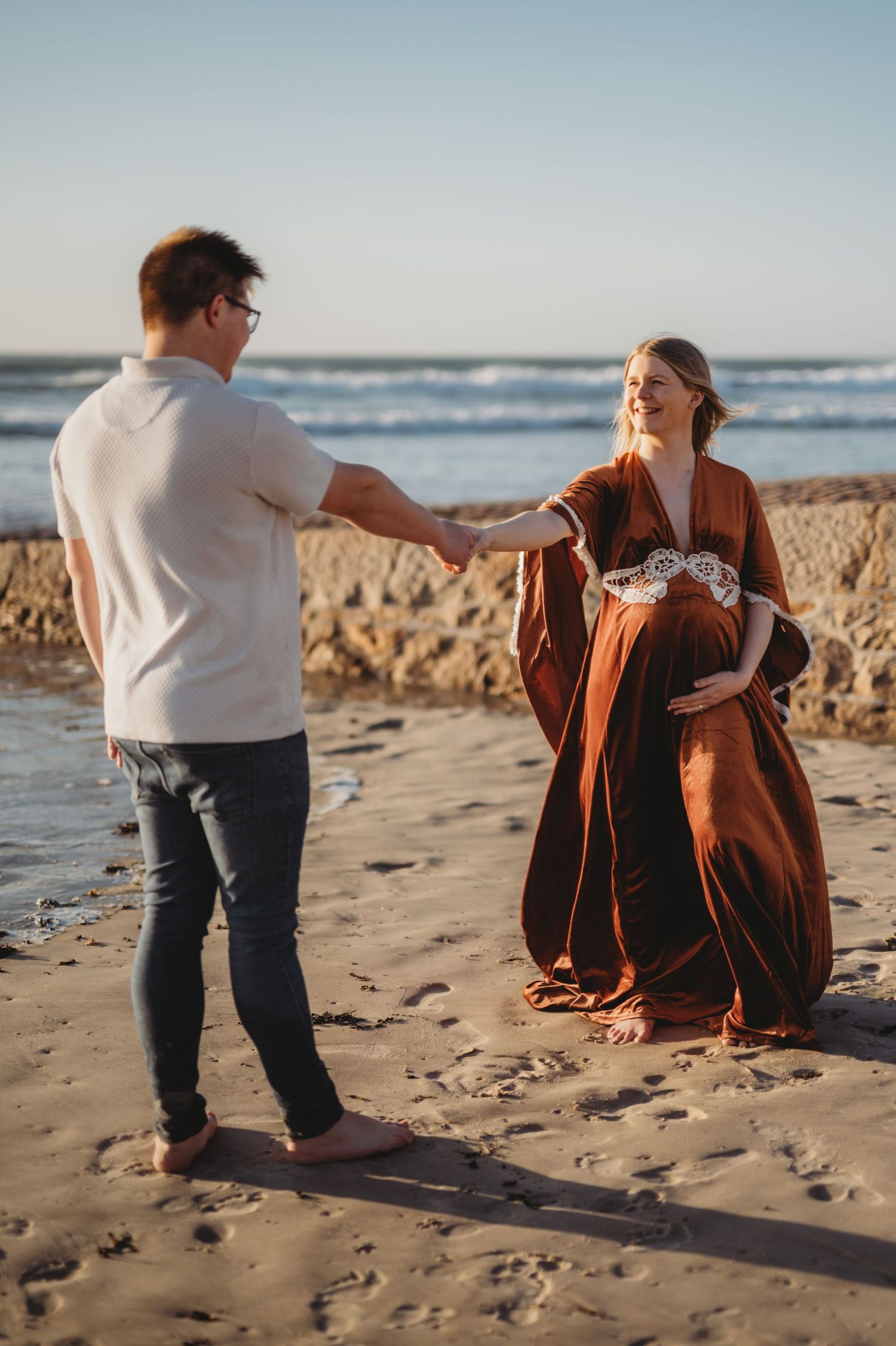 Couple holding hands on sunny beach, pregnant woman in rust boho dress smiling at partner.