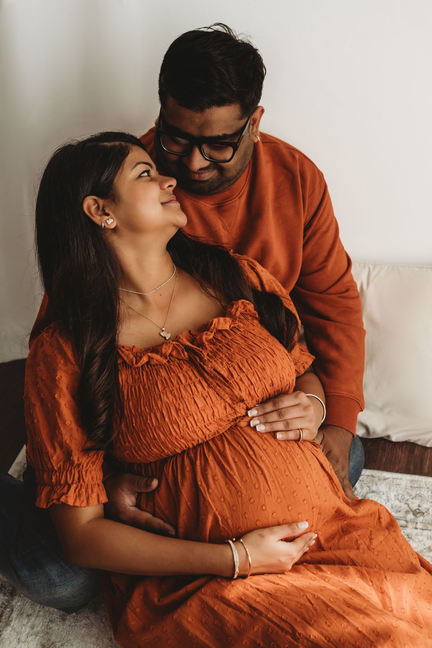 An expecting couple in matching rust outfits sit together, tenderly cradling her pregnant belly.