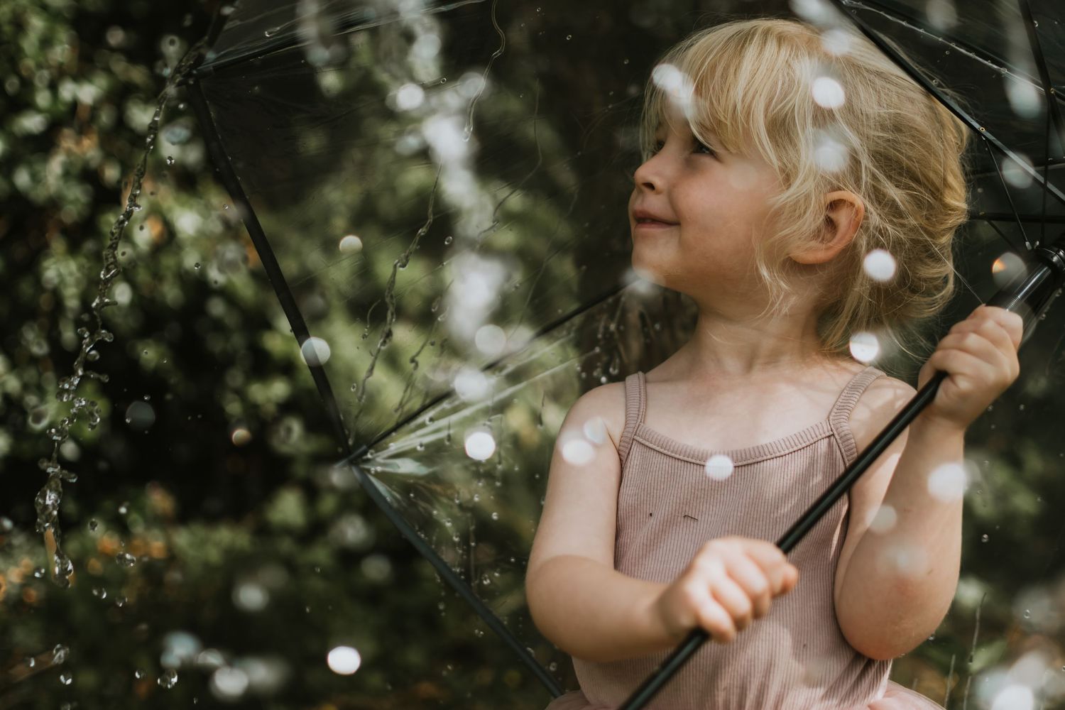 A young blonde girl holds a clear umbrella outdoors, smiling softly amid bokeh rain drops.