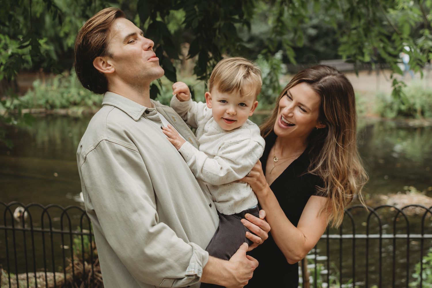 Young couple laughing outdoors in a park while holding their happy toddler child.