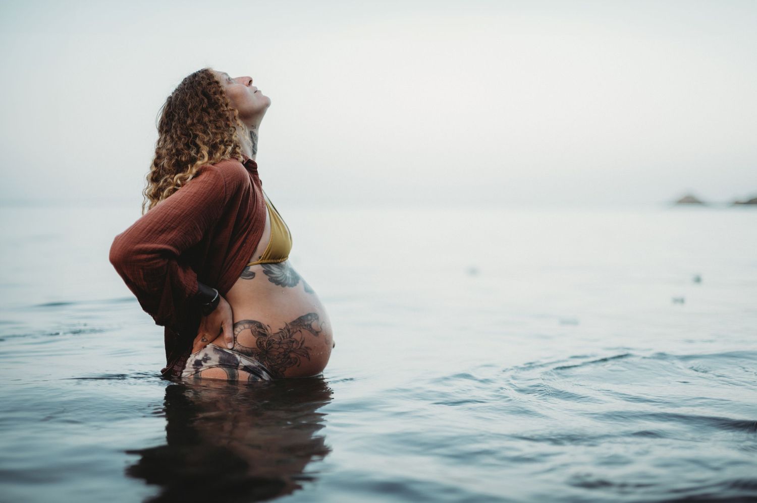 Pregnant woman standing in calm ocean water, wearing a rust top, with curly hair and head tilted back peacefully.