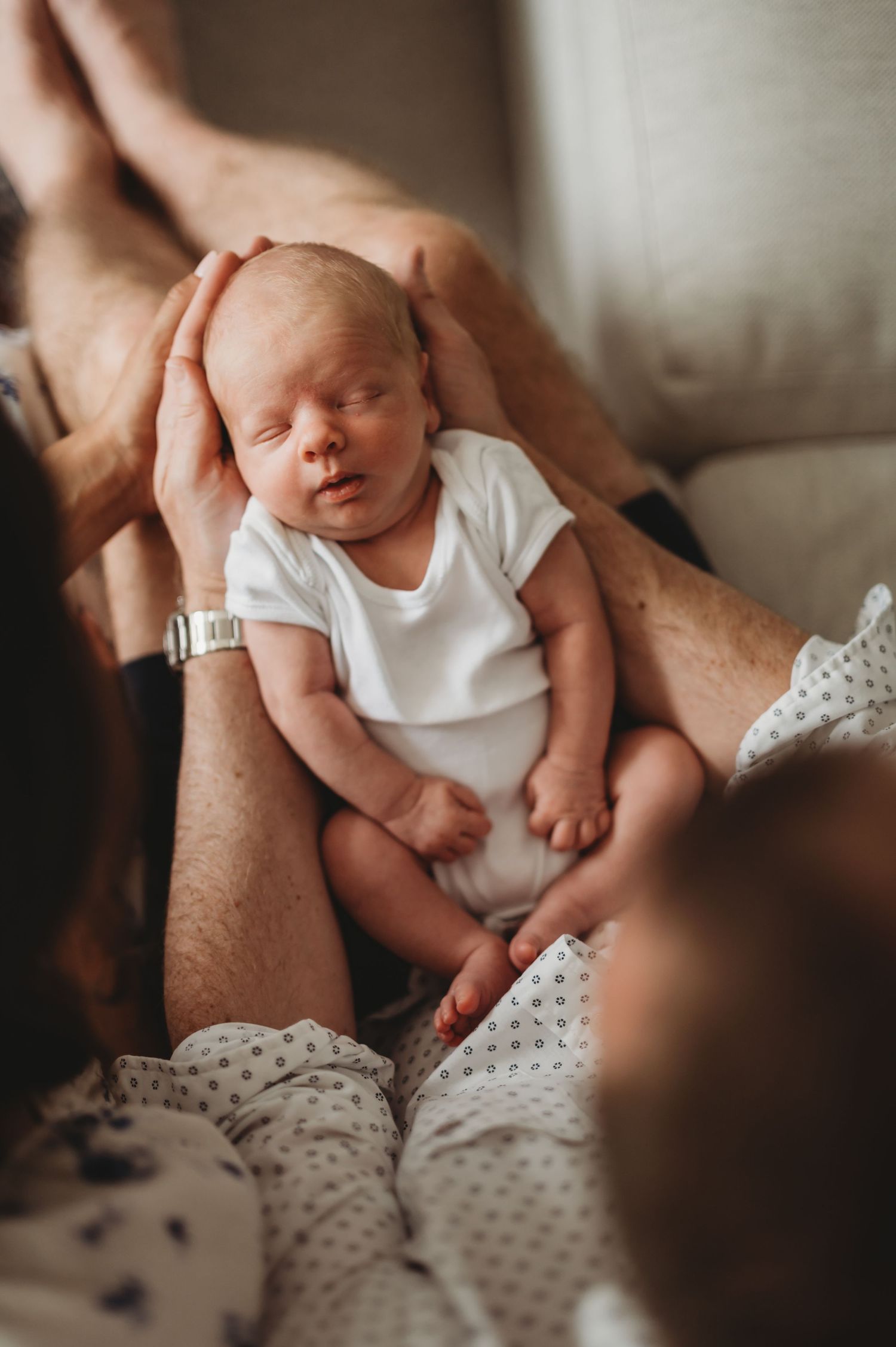 Newborn baby in white onesie sleeping peacefully while parents cradle and support the infant with gentle hands.