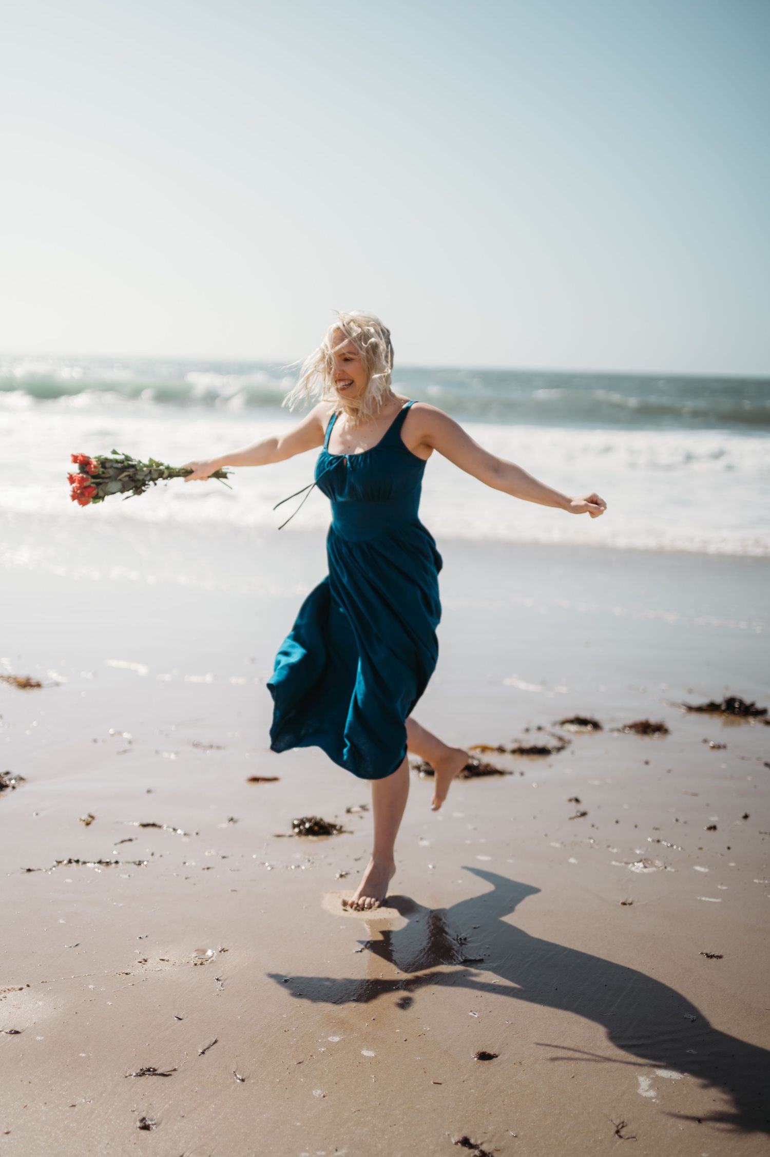 Blonde woman in teal dress dancing on beach holding red roses with arms outstretched.