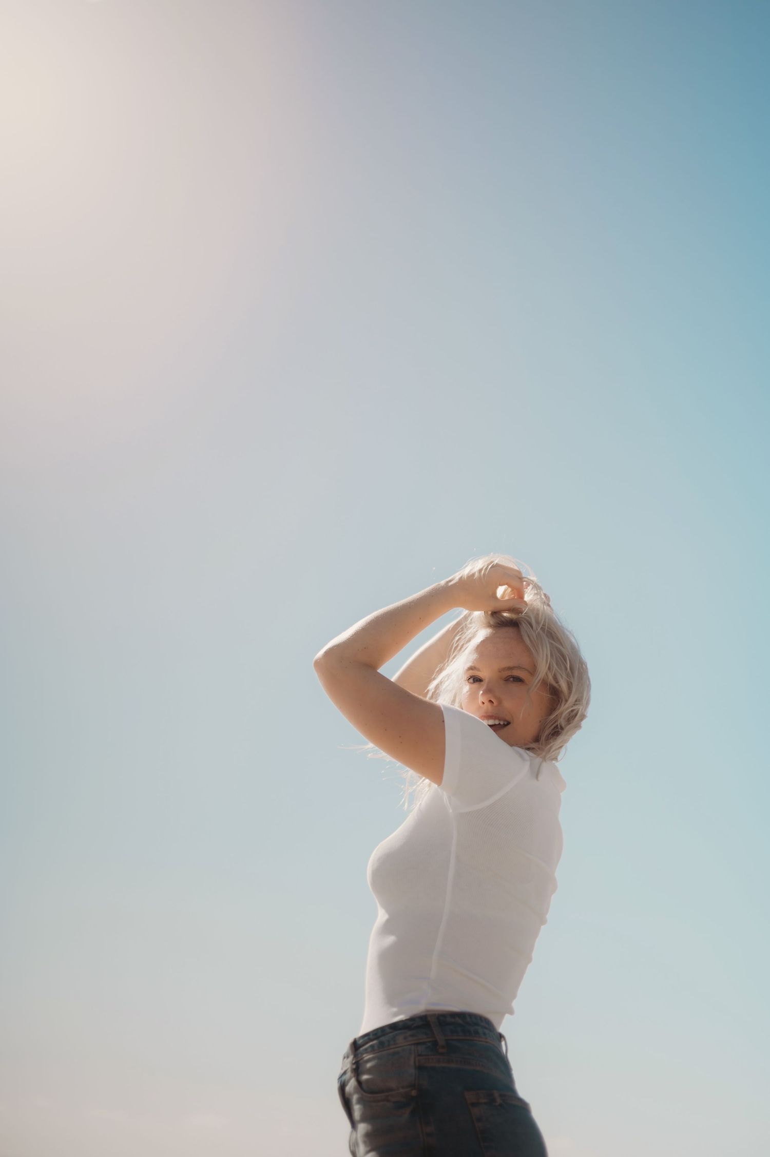 Blonde woman in white top posing against a bright blue sky with hands raised to her head.