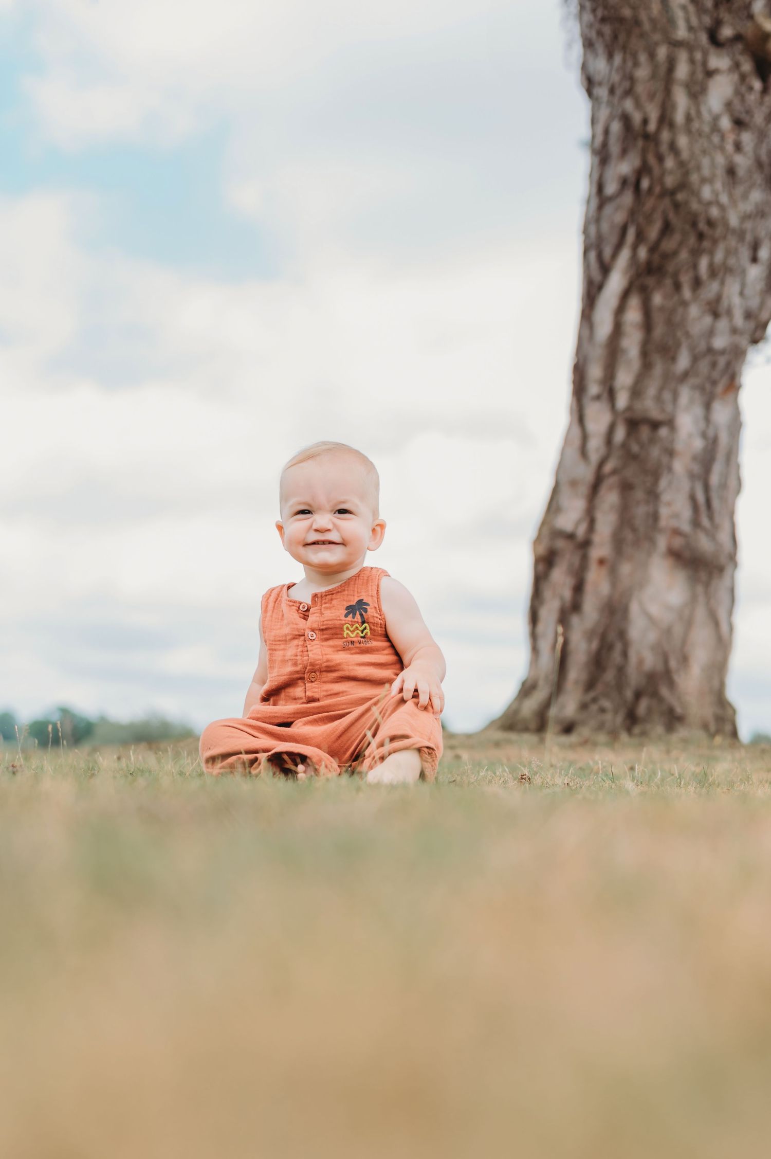 A smiling baby in an orange romper sits on grass beside a large tree under a cloudy sky.