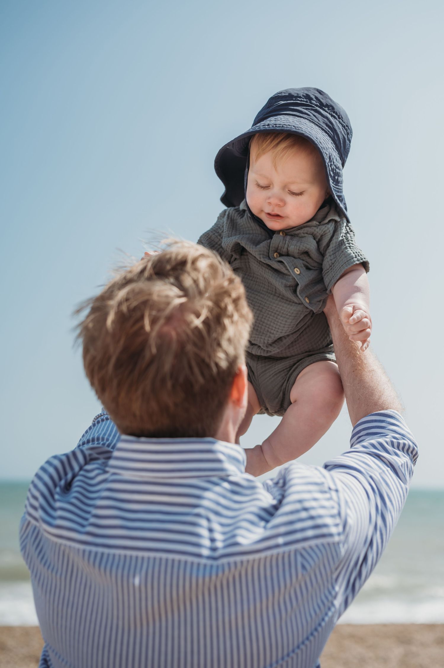 Father lifting smiling baby wearing hat at sunny beach.