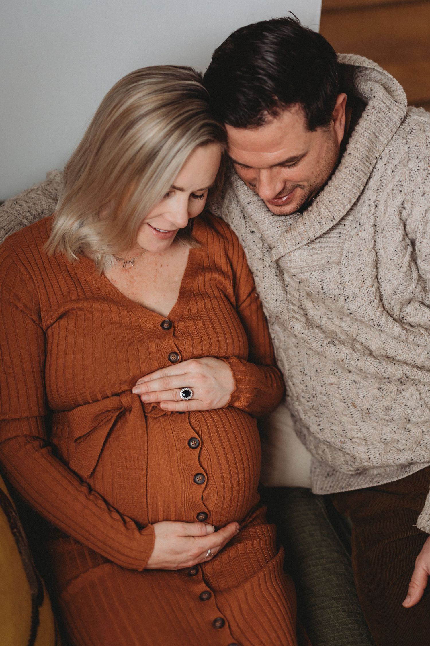 Couple in warm knitwear lovingly gazing at pregnant belly during an intimate indoor maternity photo session.